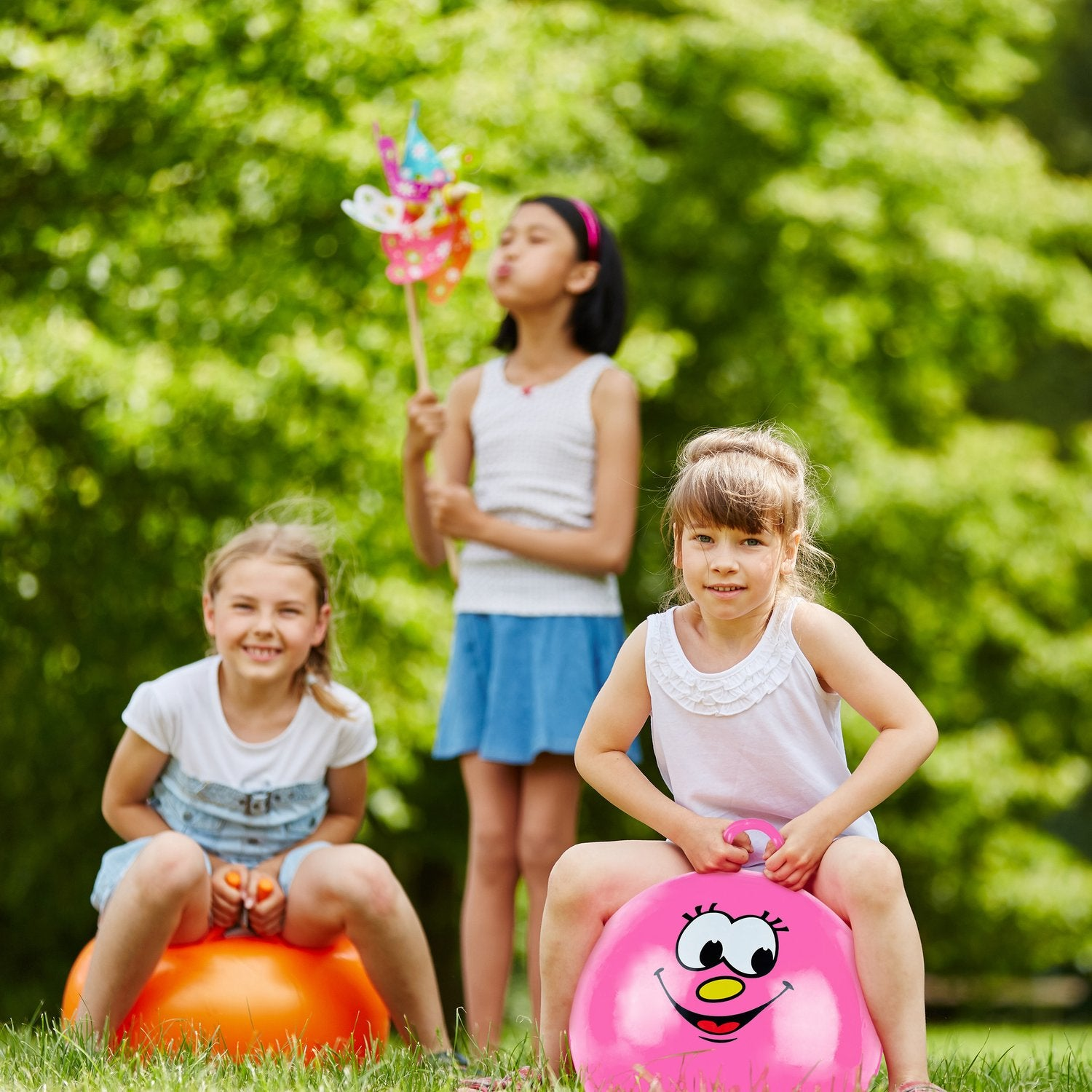 Tres niñas juegan al aire libre con pelotas saltarinas y un molinillo de viento.