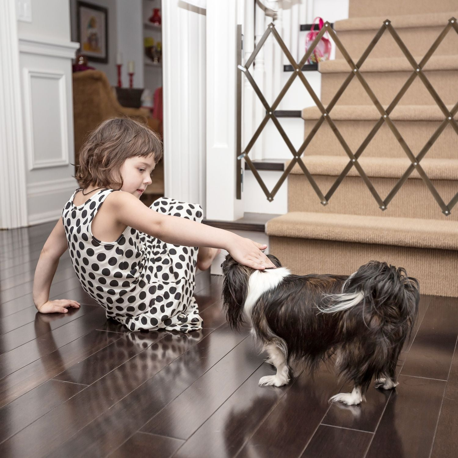 Una niña con vestido de lunares acaricia a un perro en el suelo oscuro cerca de una escalera.