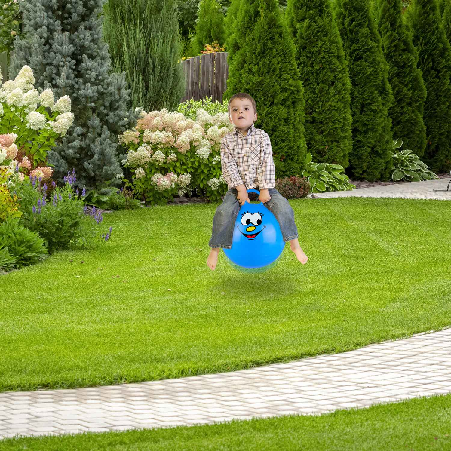 Un niño pequeño salta sobre una pelota azul con cara en un jardín verde exuberante con árboles.