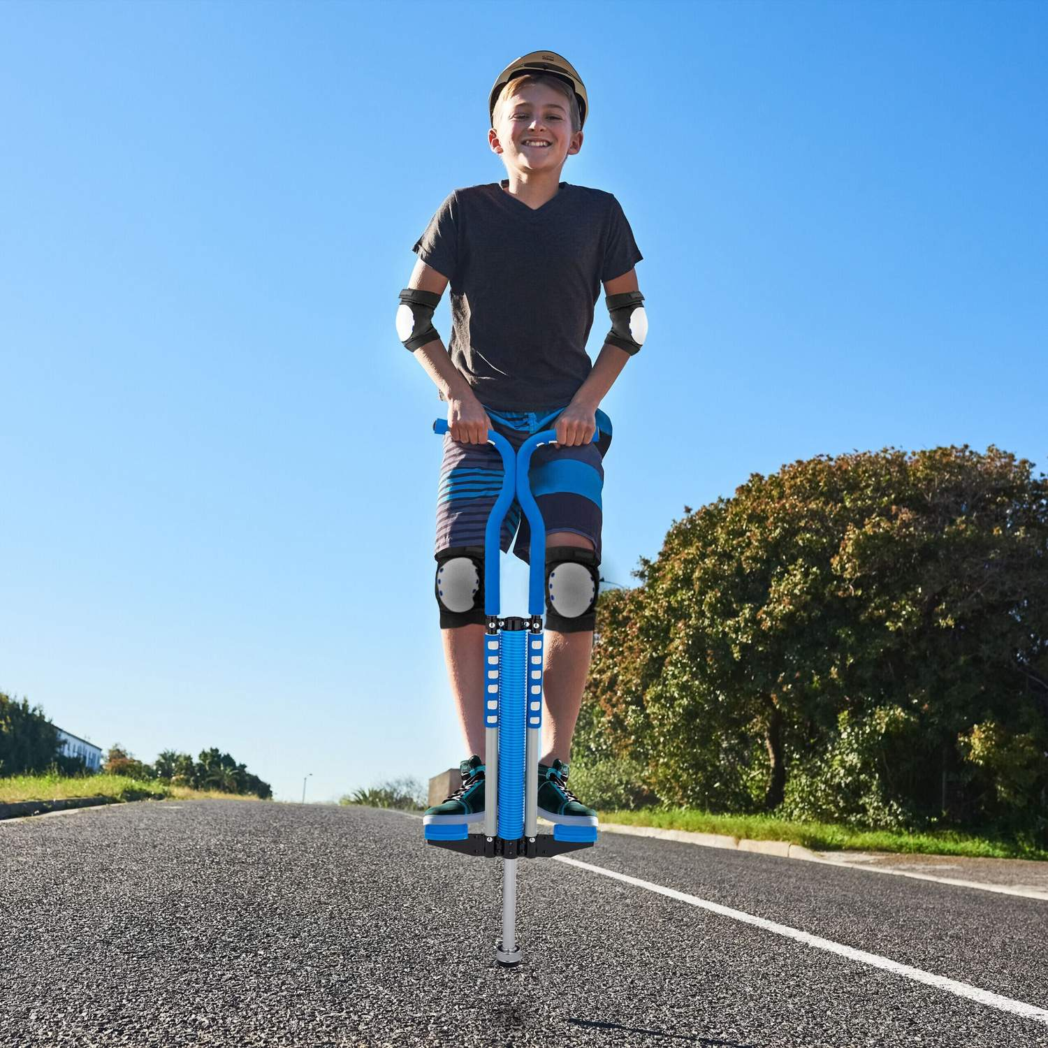 Un niño sonriente con casco y equipo de protección se para en un pogo azul en una carretera de asfalto bajo un cielo despejado.