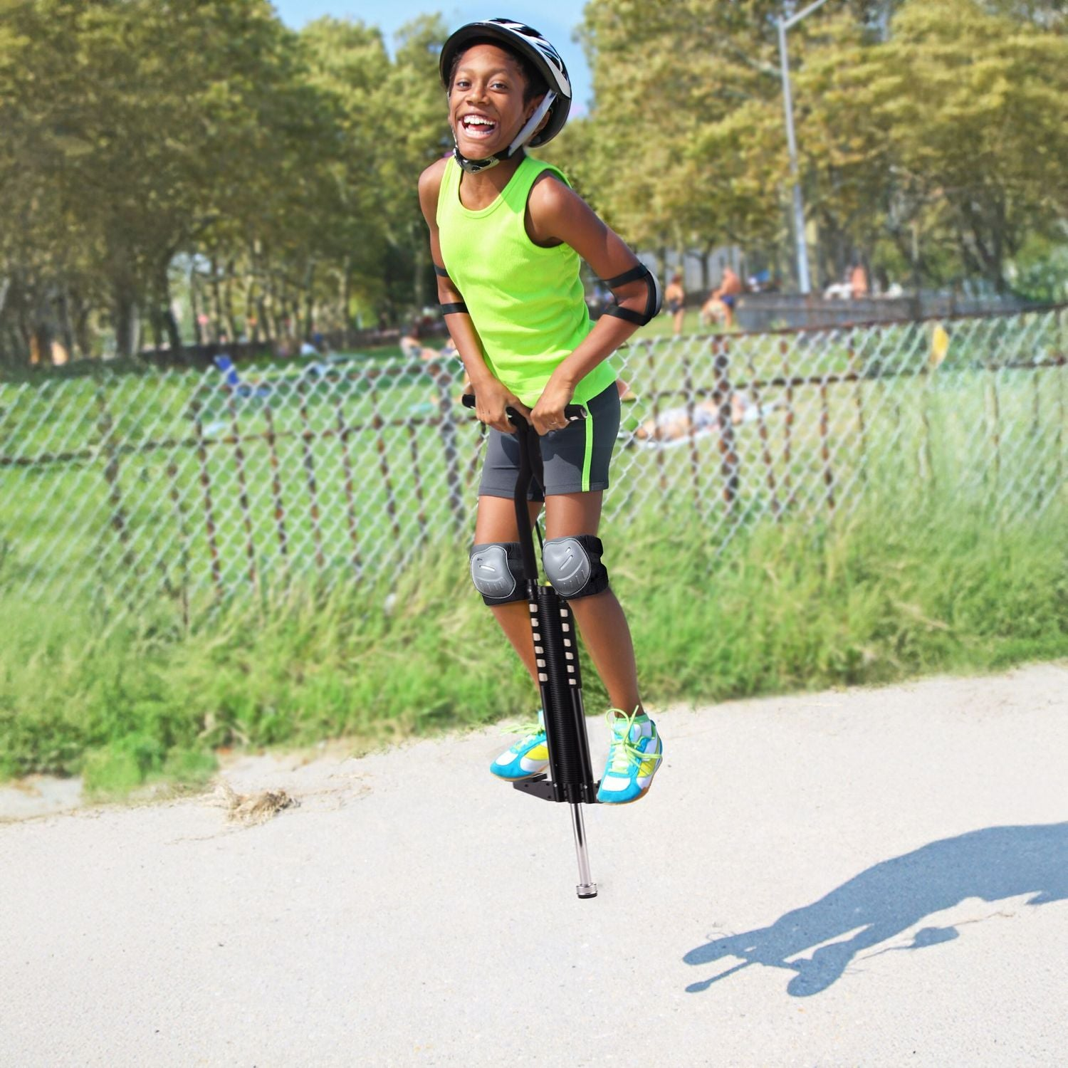 Un niño sonriente con casco y equipo de protección salta en un pogo stick en un camino pavimentado con árboles verdes al fondo.