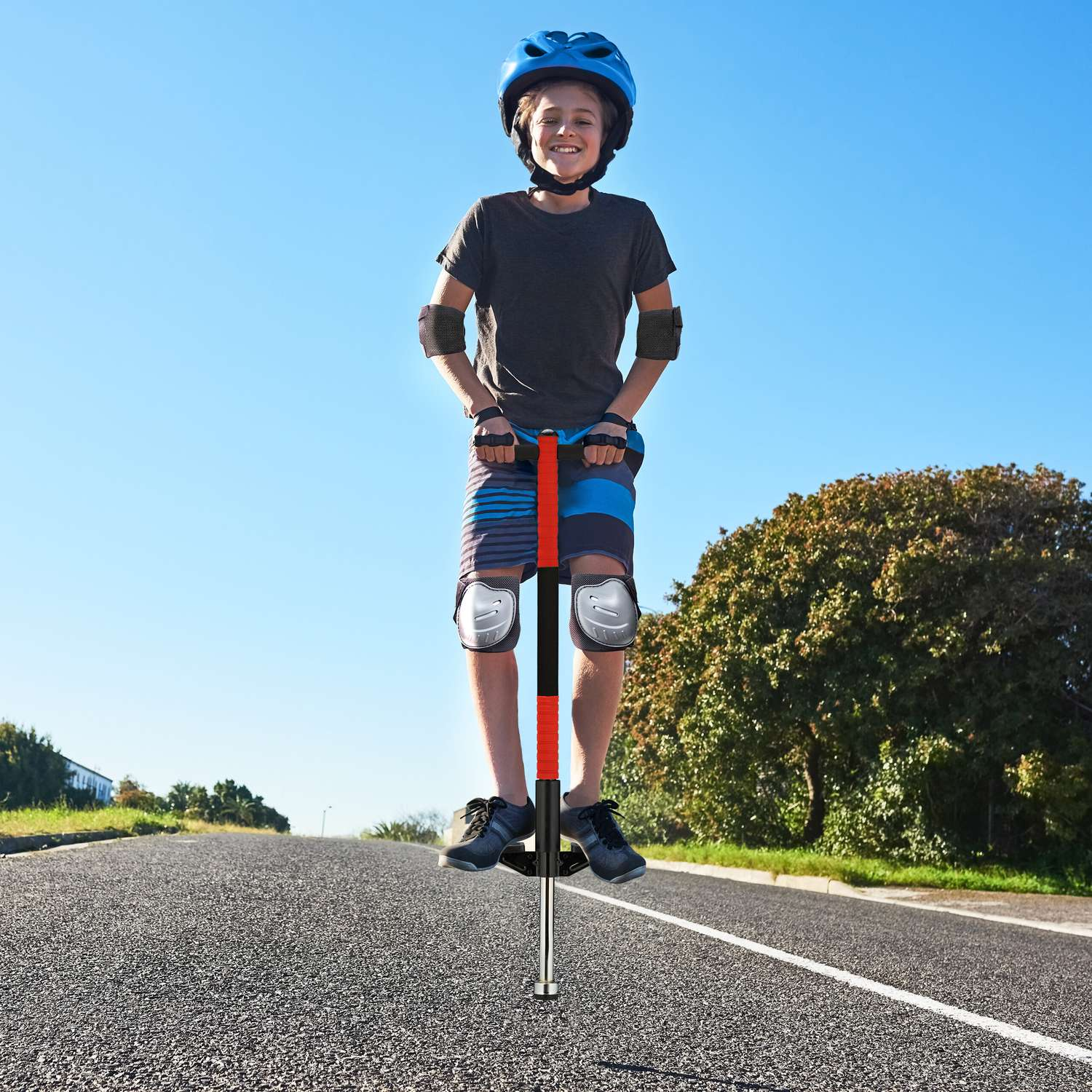 Un niño con casco y protecciones salta en un pogo stick rojo y negro en una carretera.