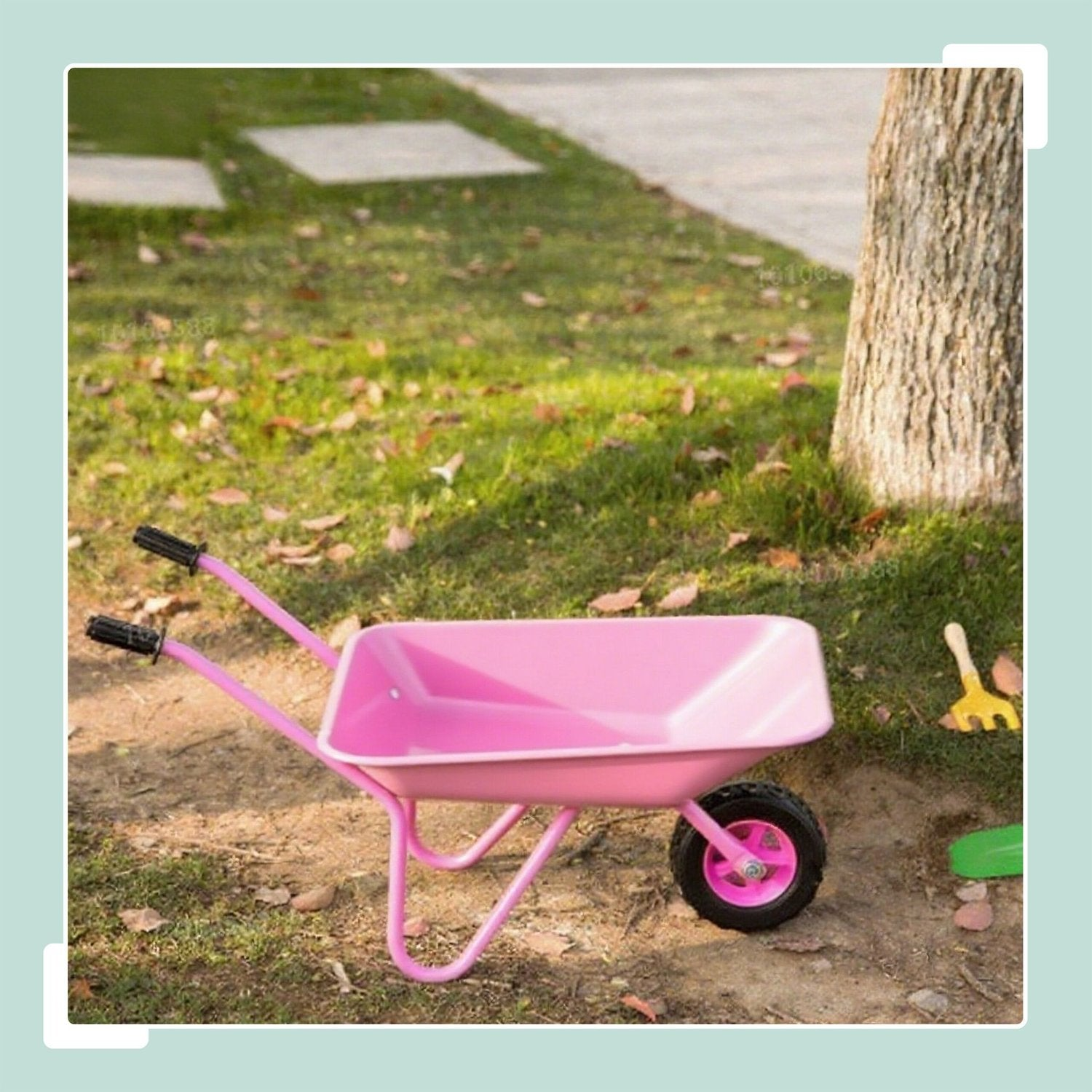 Una carretilla rosa infantil está en el césped junto a un árbol con juguetes.