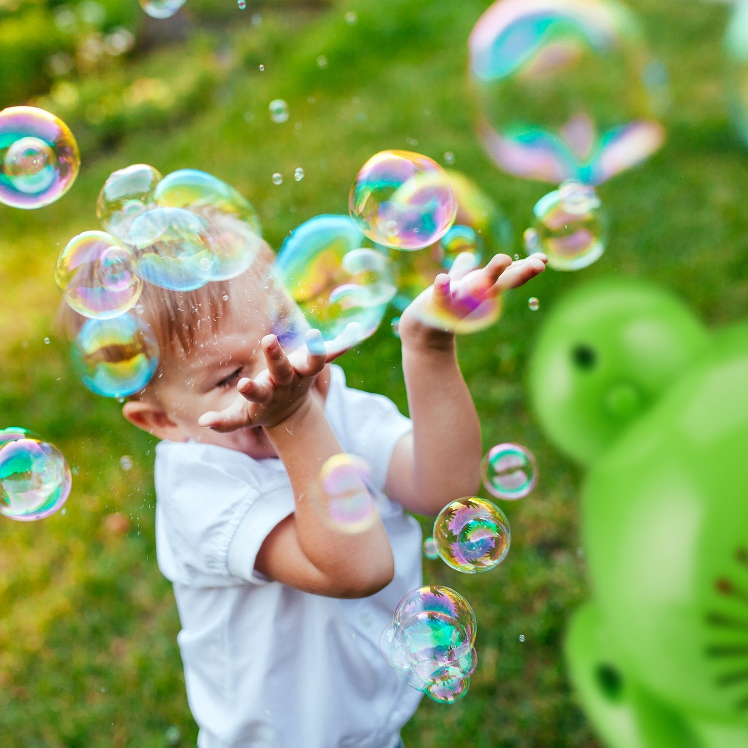Un niño pequeño intenta atrapar burbujas de jabón en un patio trasero verde.