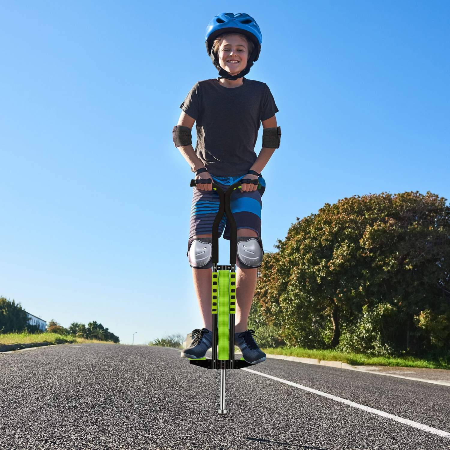 Niño con casco en pogo stick verde en carretera con árboles detrás.