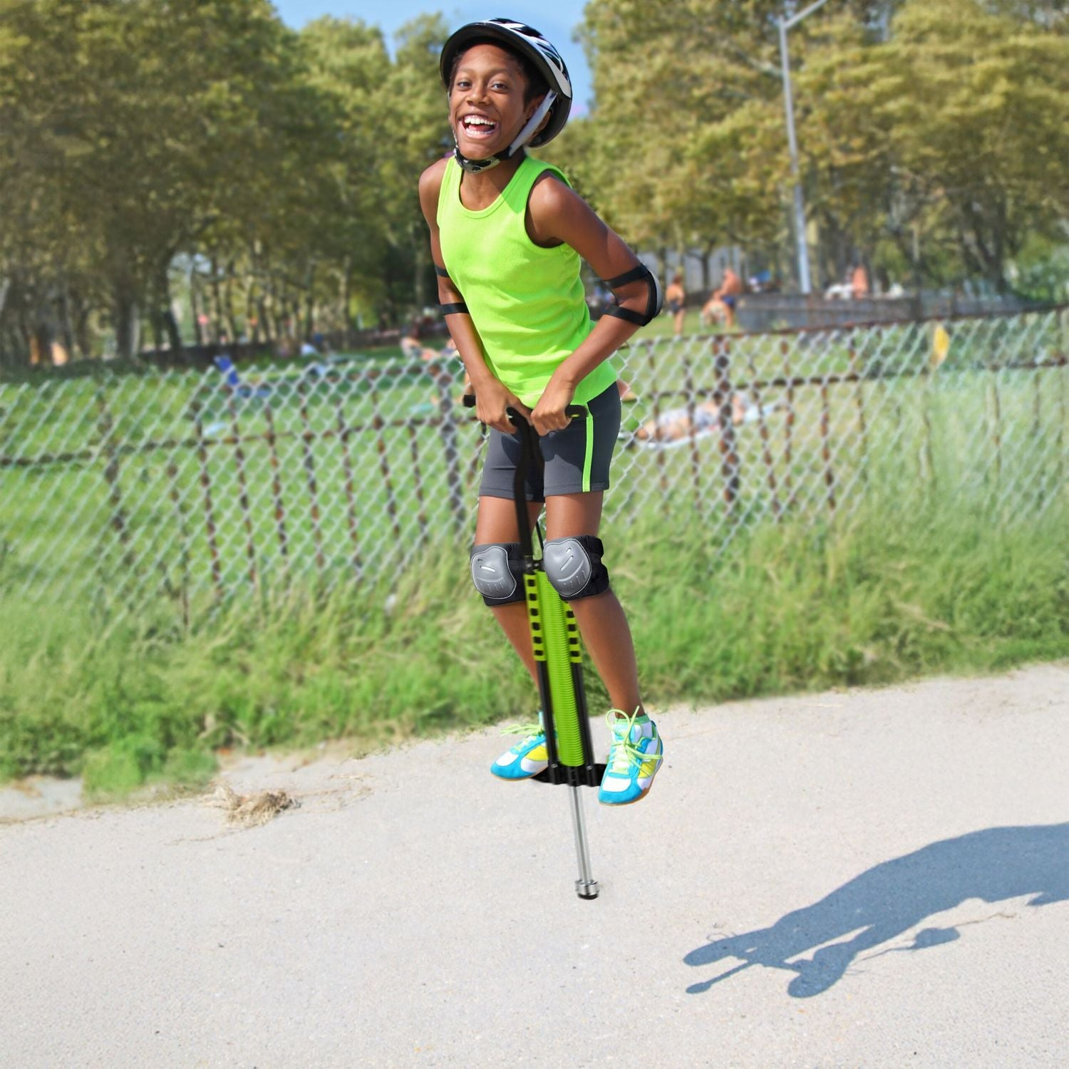 Un niño sonriente con casco y coderas salta alto en un pogo verde en un camino pavimentado con árboles y una valla al fondo.