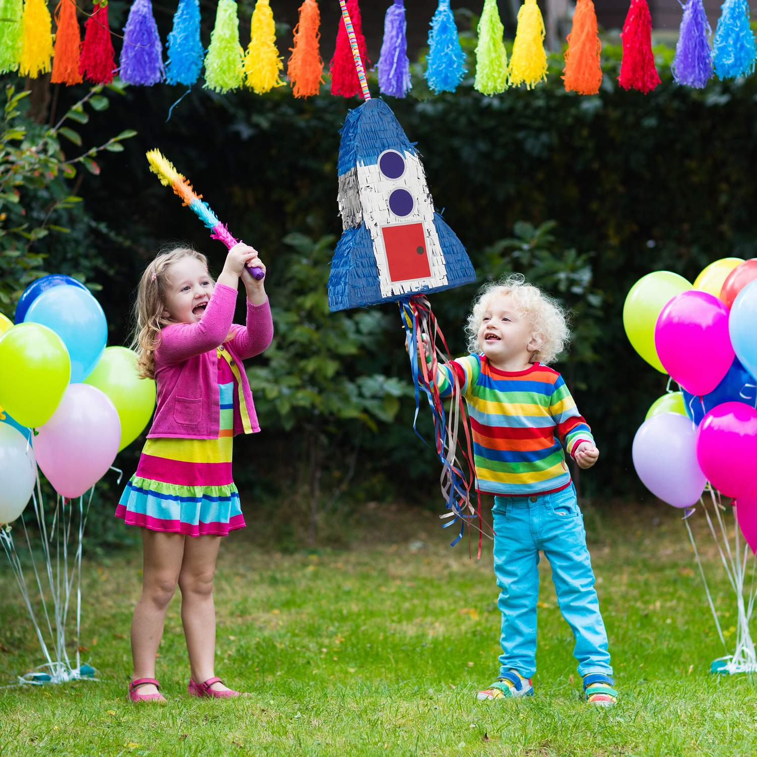 Dos niños golpean una piñata de cohete en una fiesta en el jardín con globos.