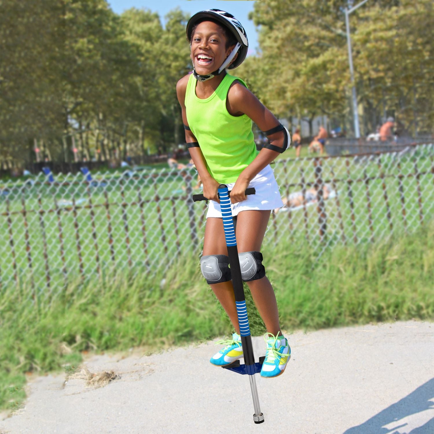 Una niña de verde salta en un pogo stick azul en un parque. Lleva equipo de protección.