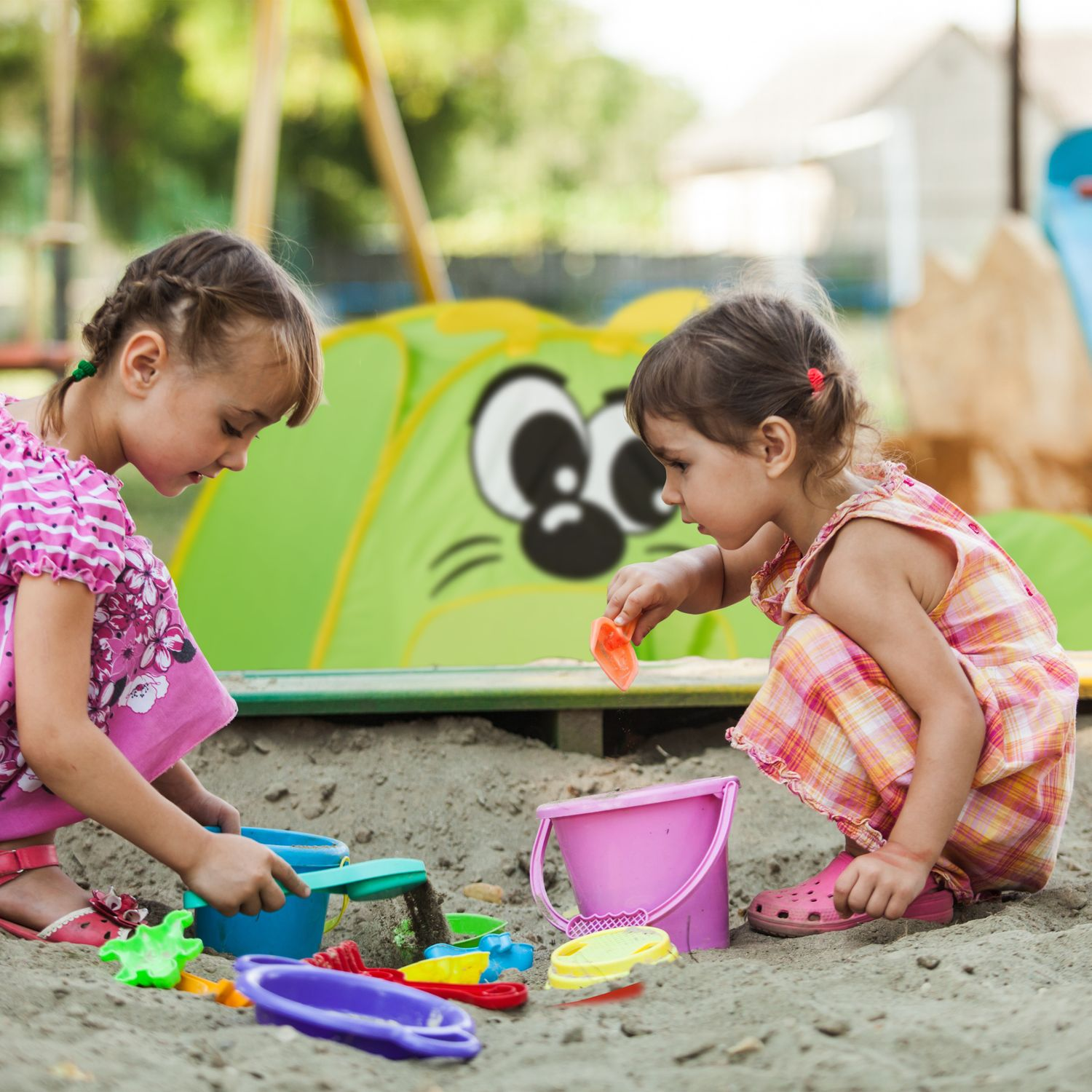 Dos niñas juegan en un arenero con juguetes coloridos y una carpa verde.