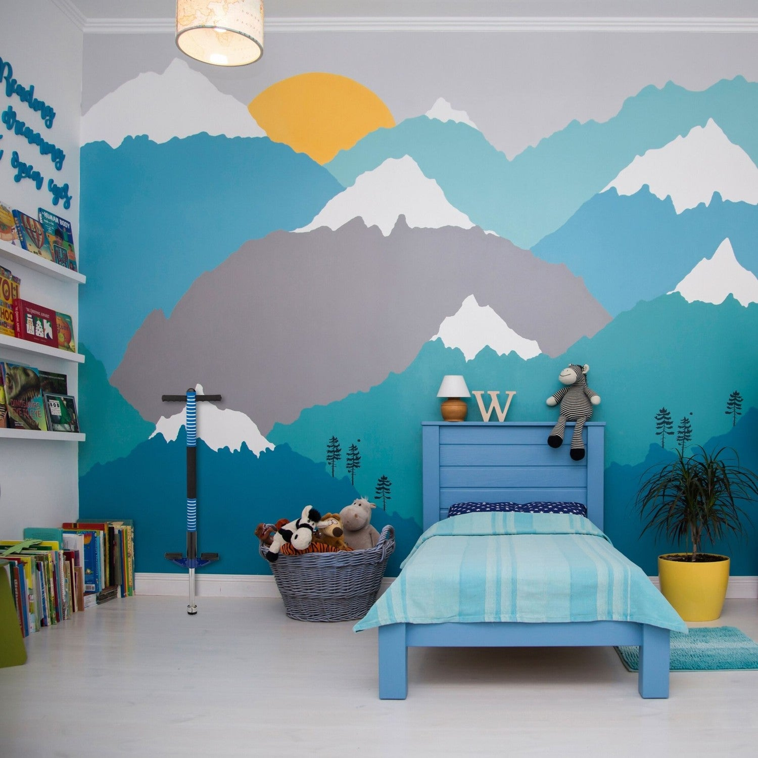 Habitación infantil con mural de montaña, cama azul, cesto de juguetes y libros.