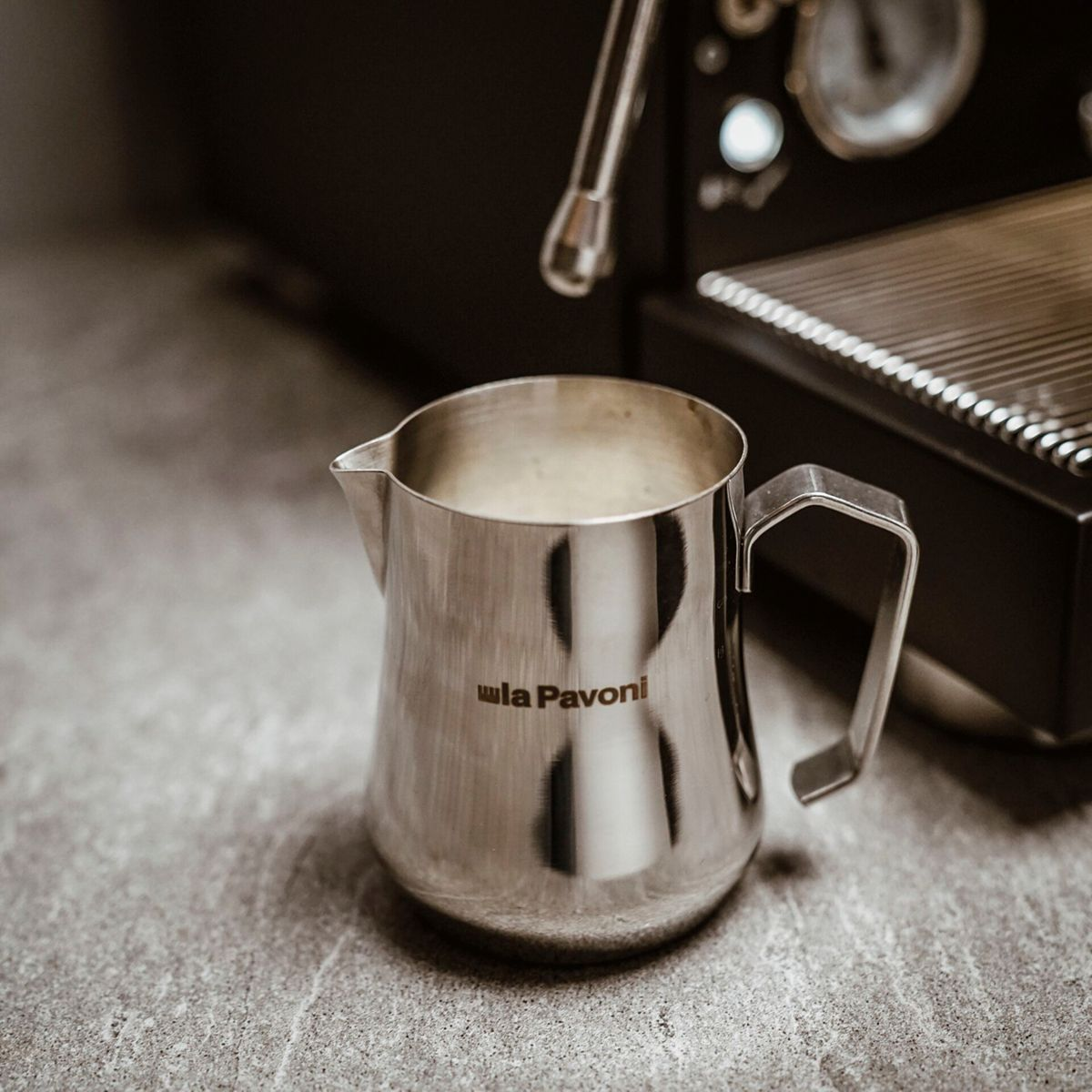 Ein silberner Milchaufschäumer mit Griff steht auf einer grauen Arbeitsplatte neben einer Espressomaschine.