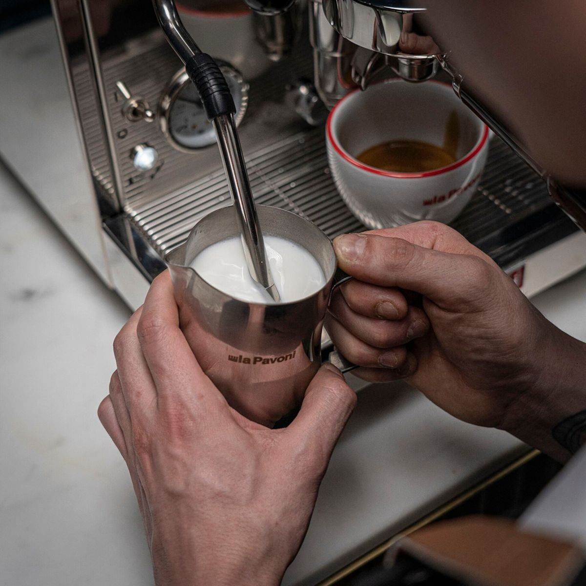 Ein Barista schäumt Milch in einem Metallkrug neben einer Espressotasse. La Pavoni Logo sichtbar.