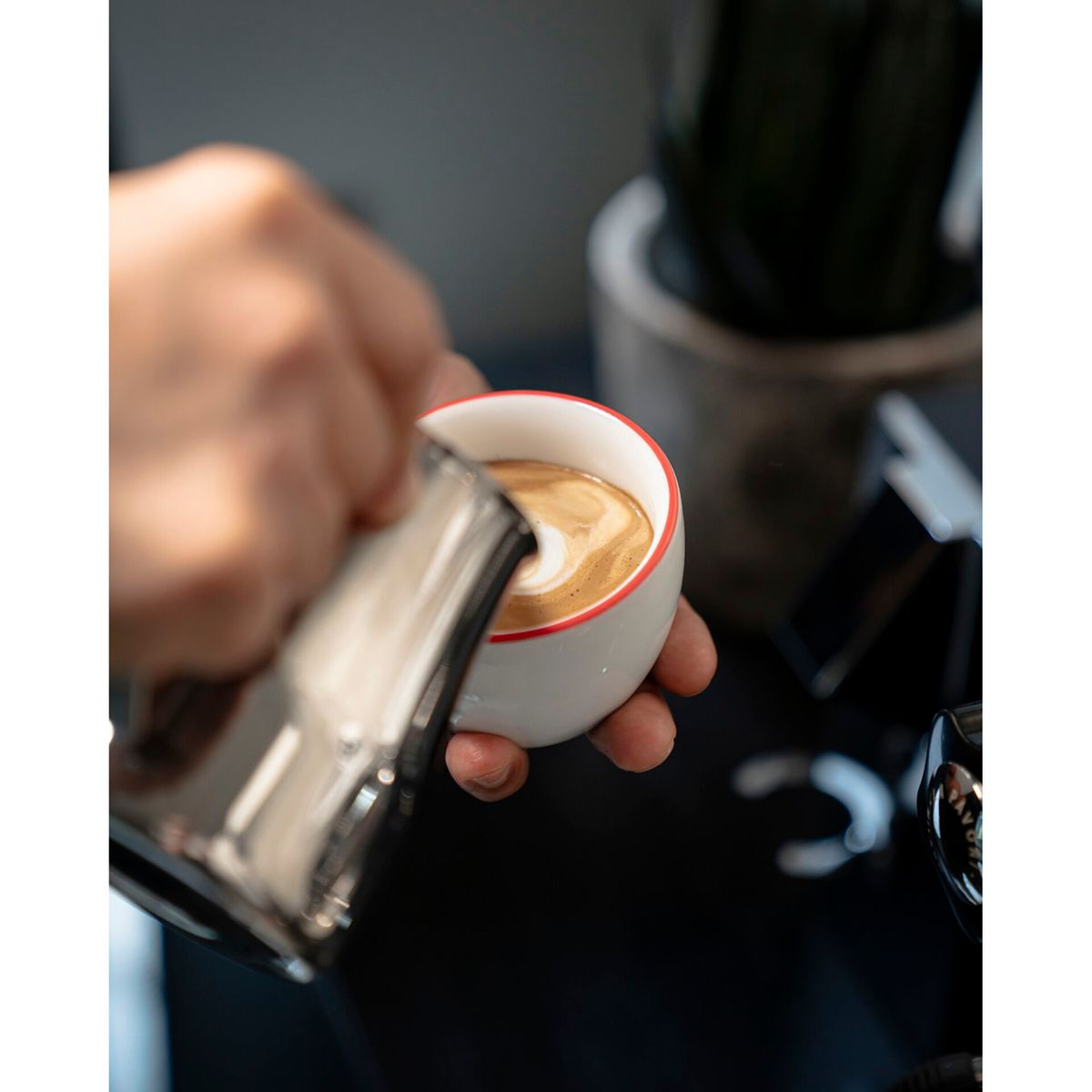 Eine Hand gießt Milch in eine weiße Kaffeetasse mit rotem Rand.
