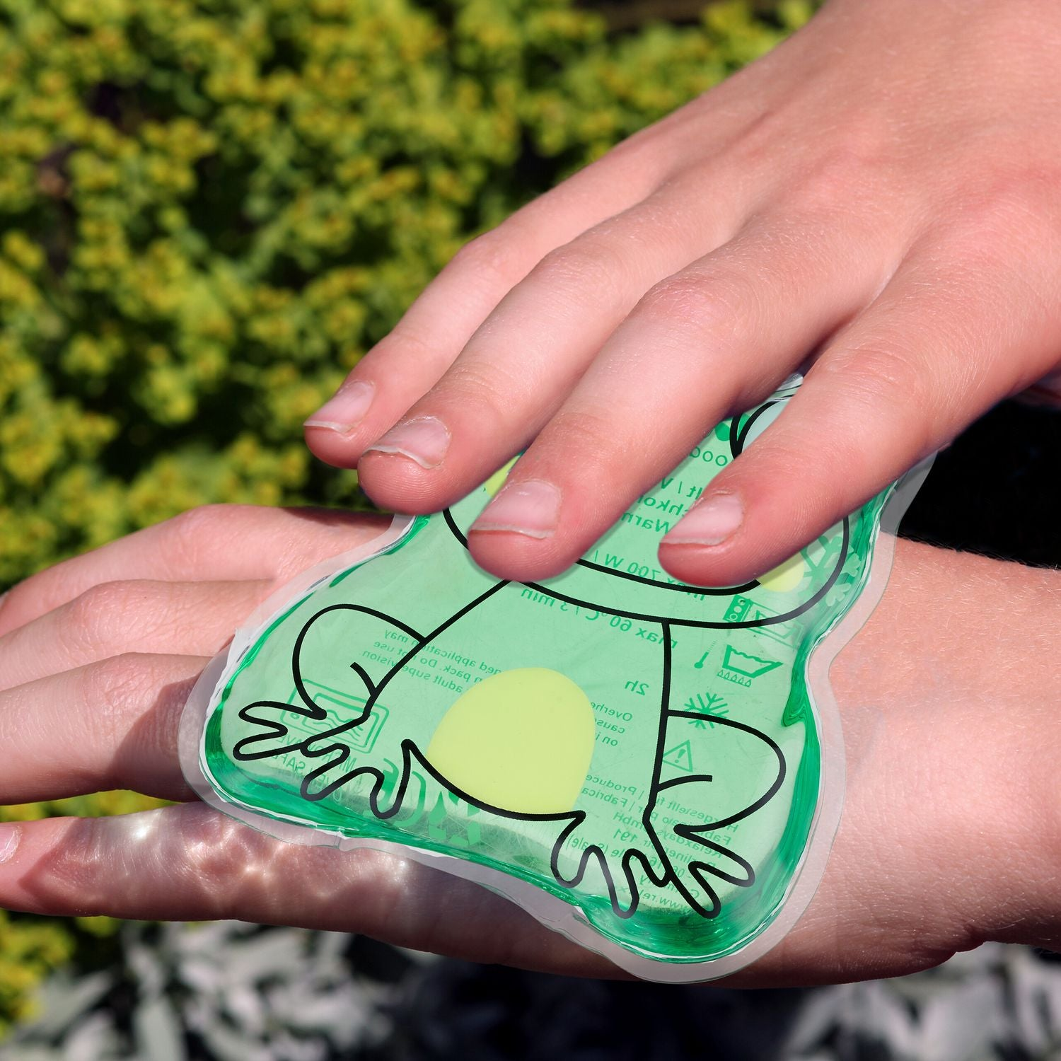 Una compresa fría verde con forma de rana en una mano, con follaje verde detrás.