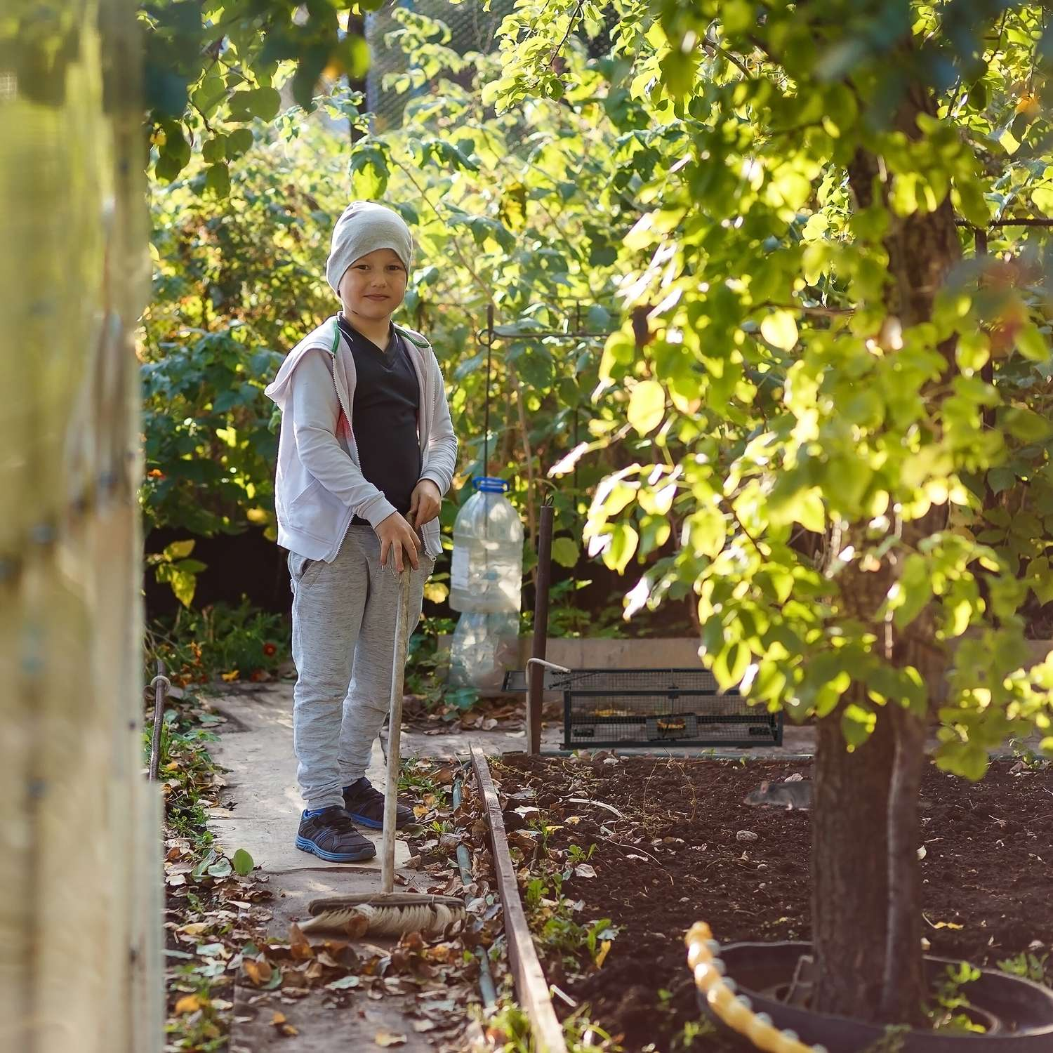 Un niño con gorro y sudadera con capucha está en el jardín con una escoba.