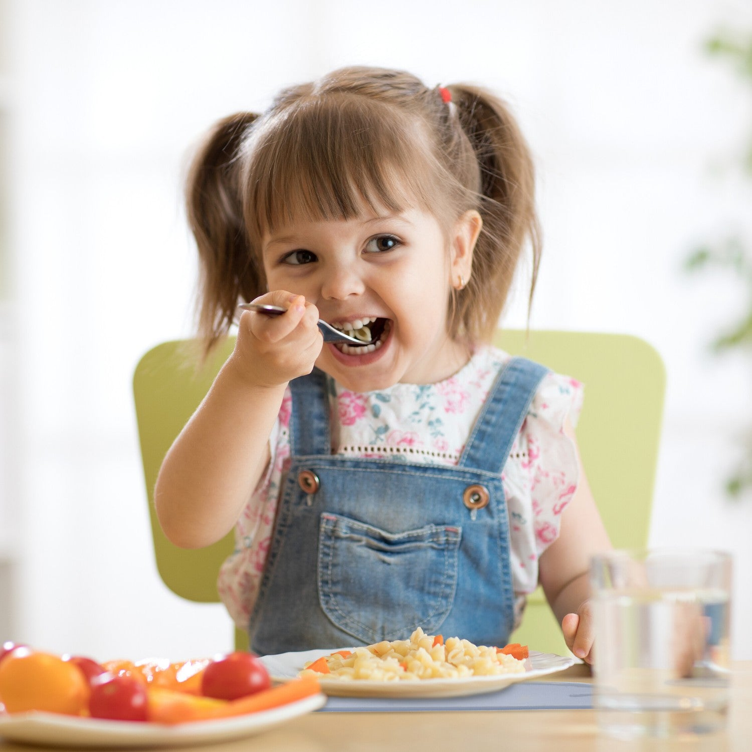 Niña pequeña come pasta con cuchara, verduras en la mesa.
