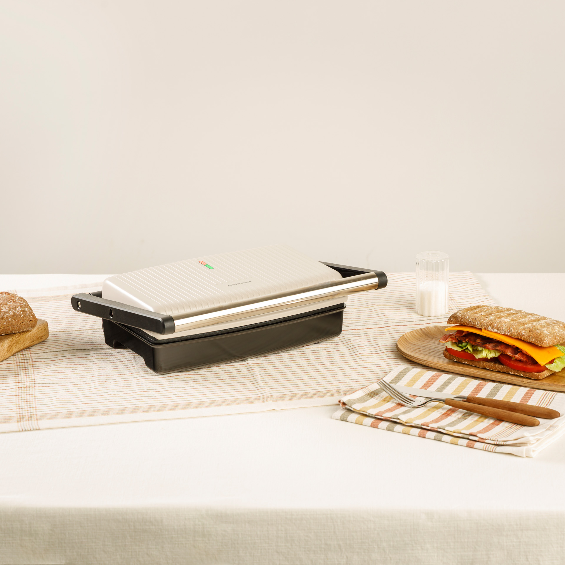 Un sandwich maker chiuso con un panino, pane e posate su un tavolo.