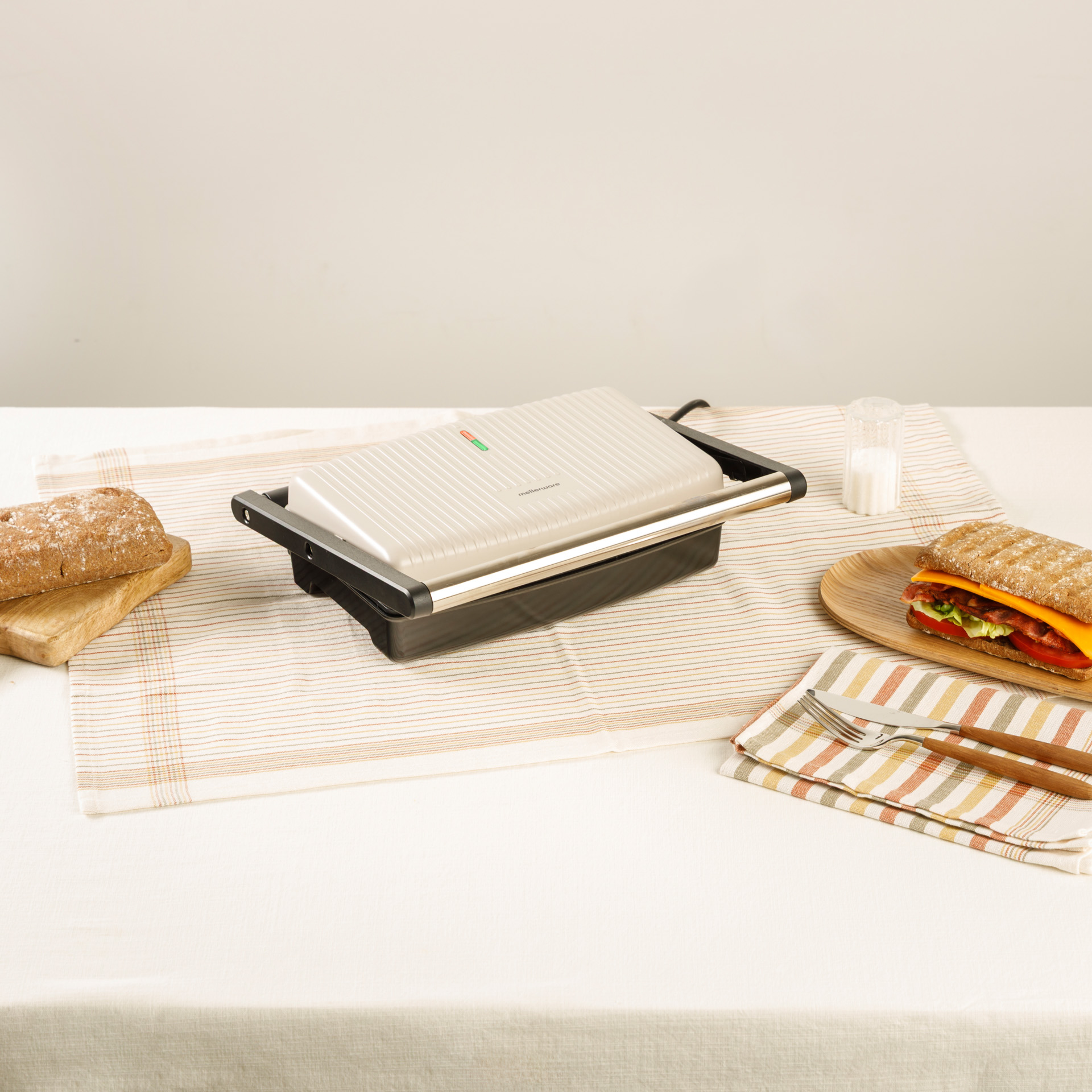 Un sandwich maker è sul tavolo con un sandwich, pane e posate.