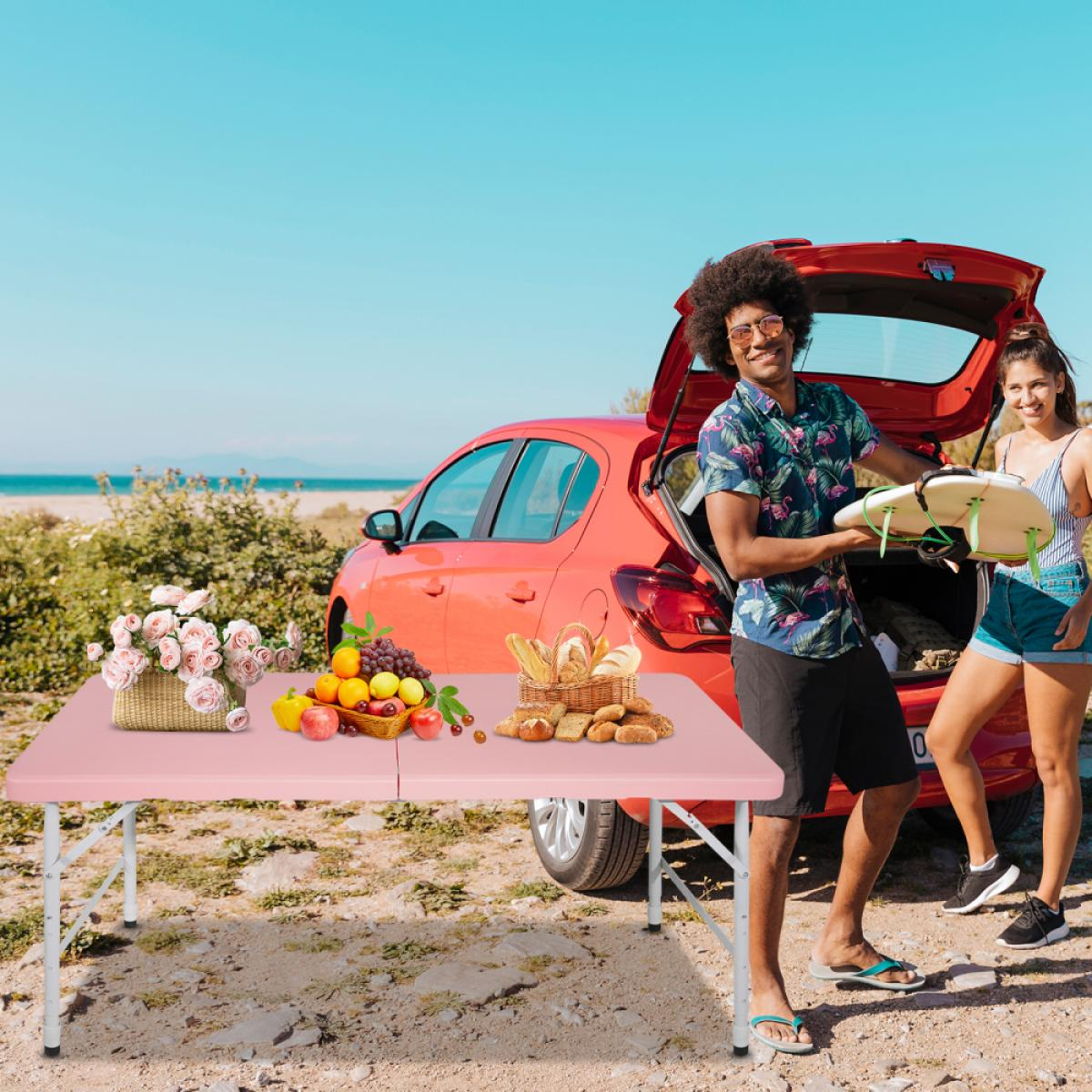 Ein Paar packt einen Surfbrett neben einem roten Auto am Strand aus, mit Picknick auf dem Tisch.