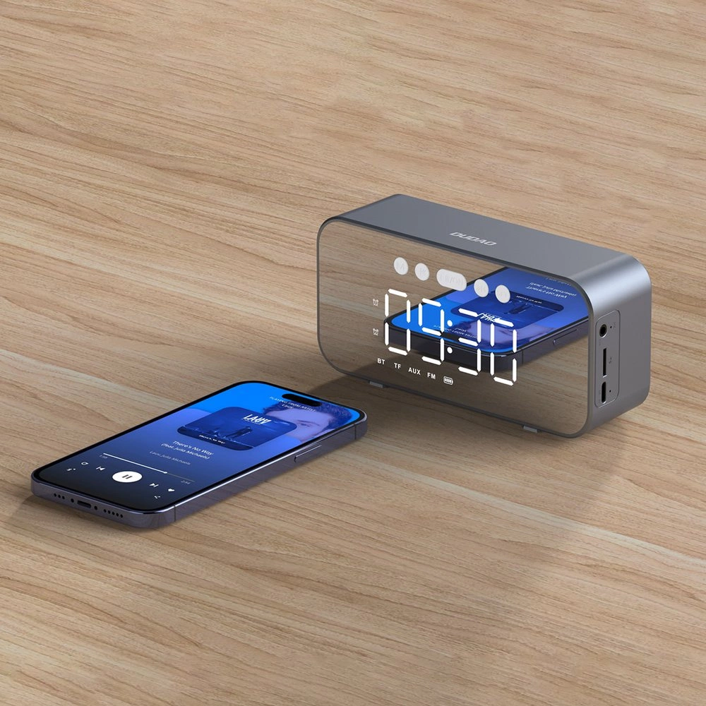 Ein grauer Lautsprecher mit Spiegel-Display und ein Smartphone liegen auf einem Holztisch.