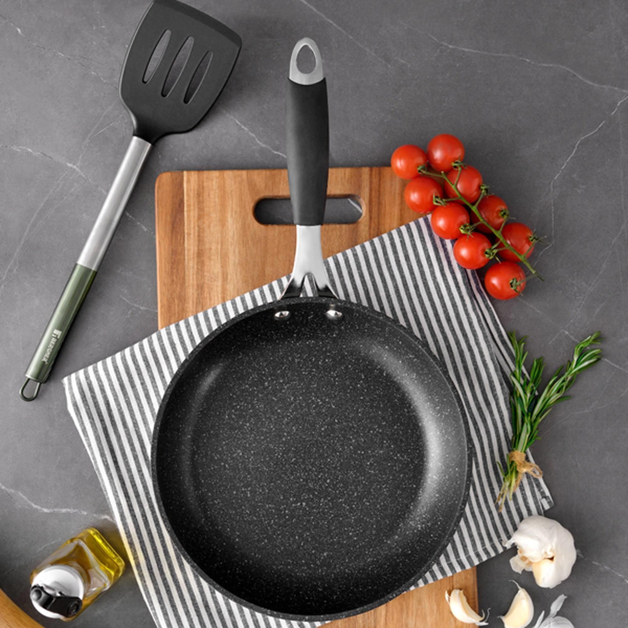 Una sartén con utensilios de cocina, tomates y ajo sobre una superficie de madera.