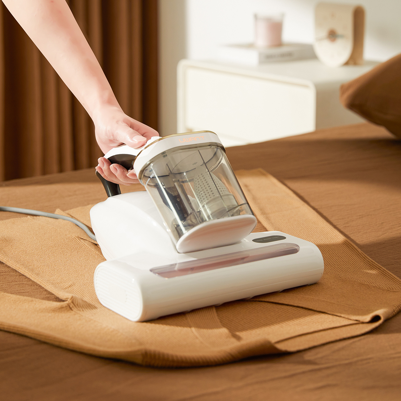 Dłoń trzyma biały odkurzacz na brązowej tkaninie.