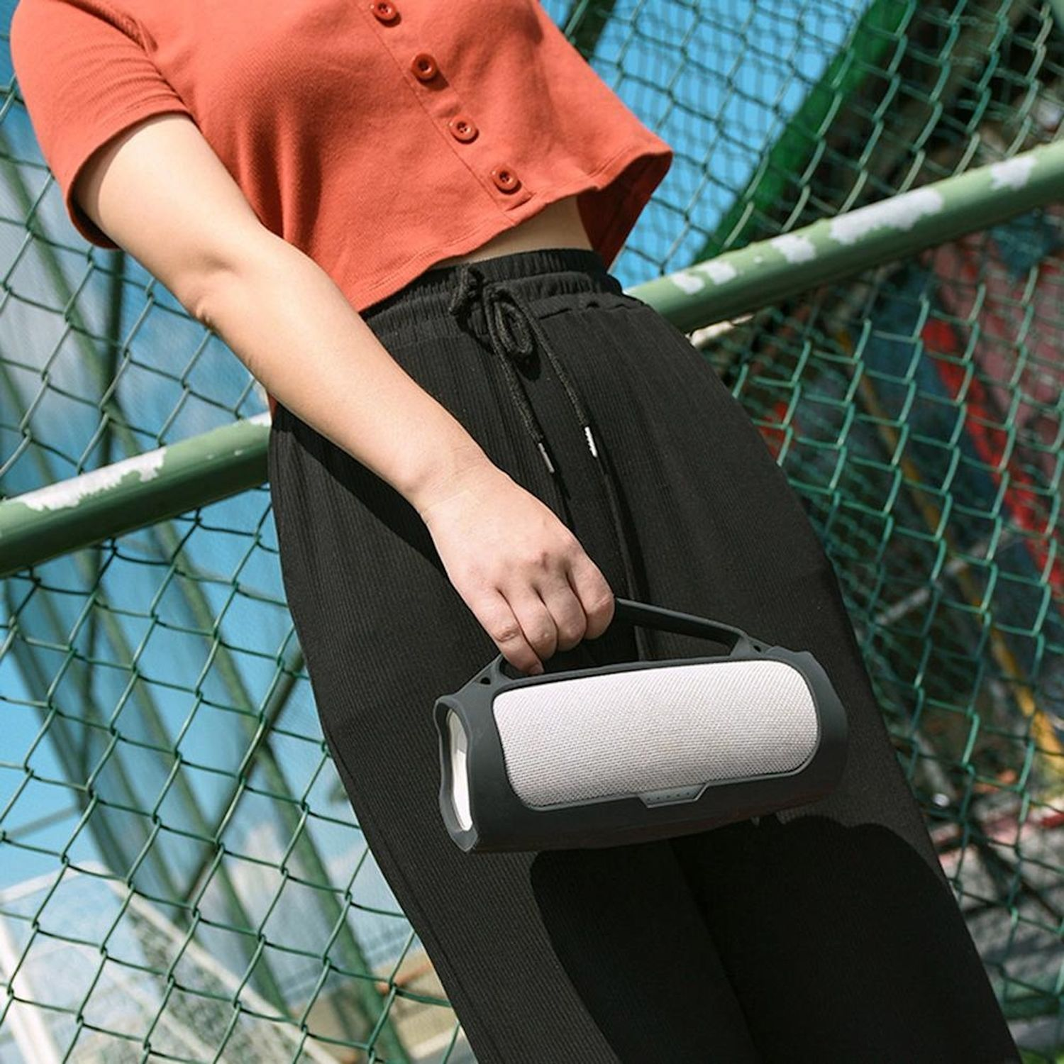 Persona con top naranja y pantalones negros sostiene un altavoz gris con asa negra.