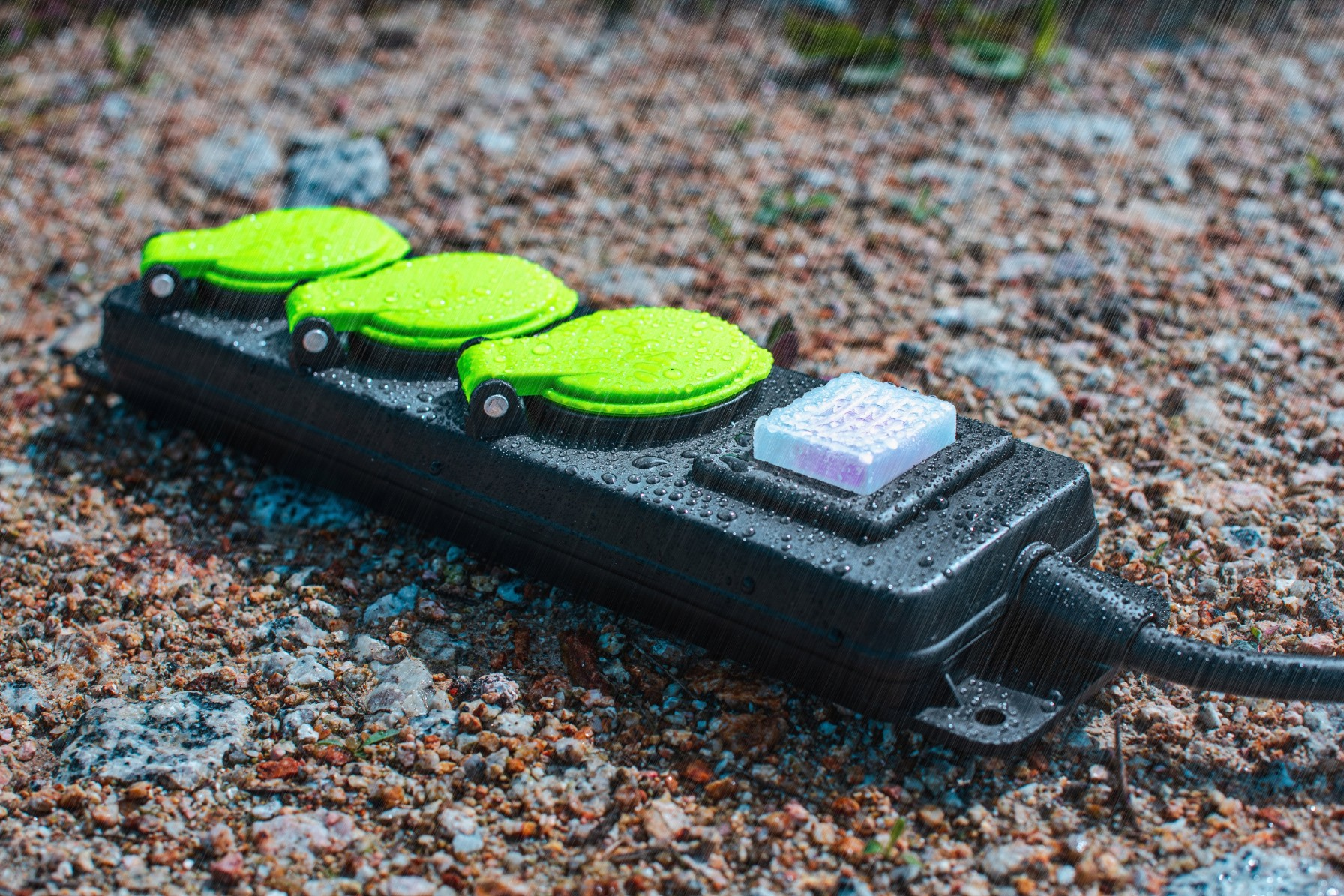 Czarna listwa zasilająca z trzema zielonymi włącznikami i niebieskim światłem, mokra od deszczu.