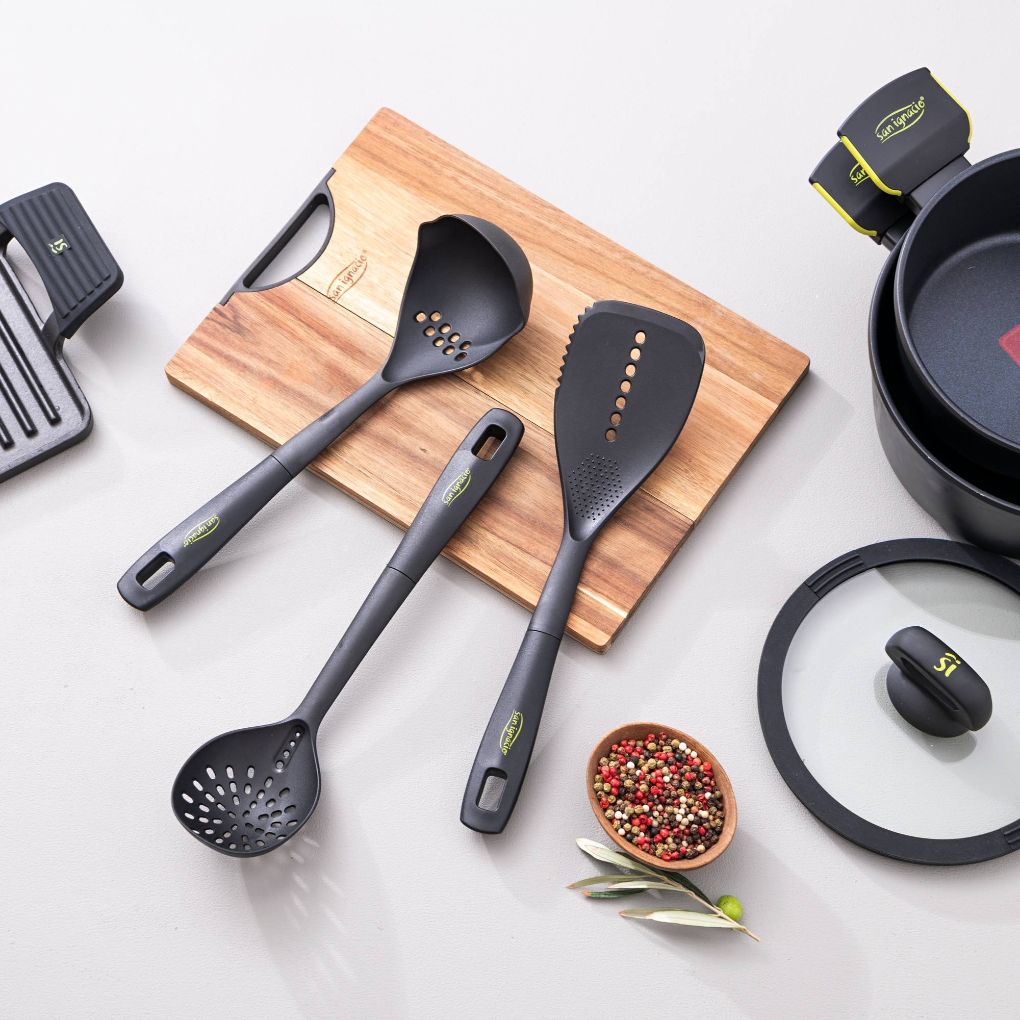 Utensilios de cocina en tabla de madera, con granos de pimienta, ramas de olivo y sartén.