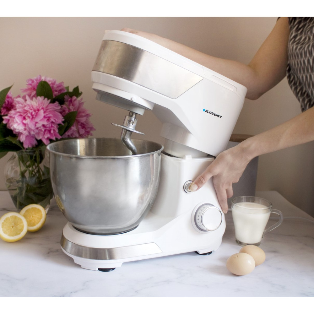 Un robot da cucina bianco con ciotola, accanto a fiori, limoni, uova e latte.