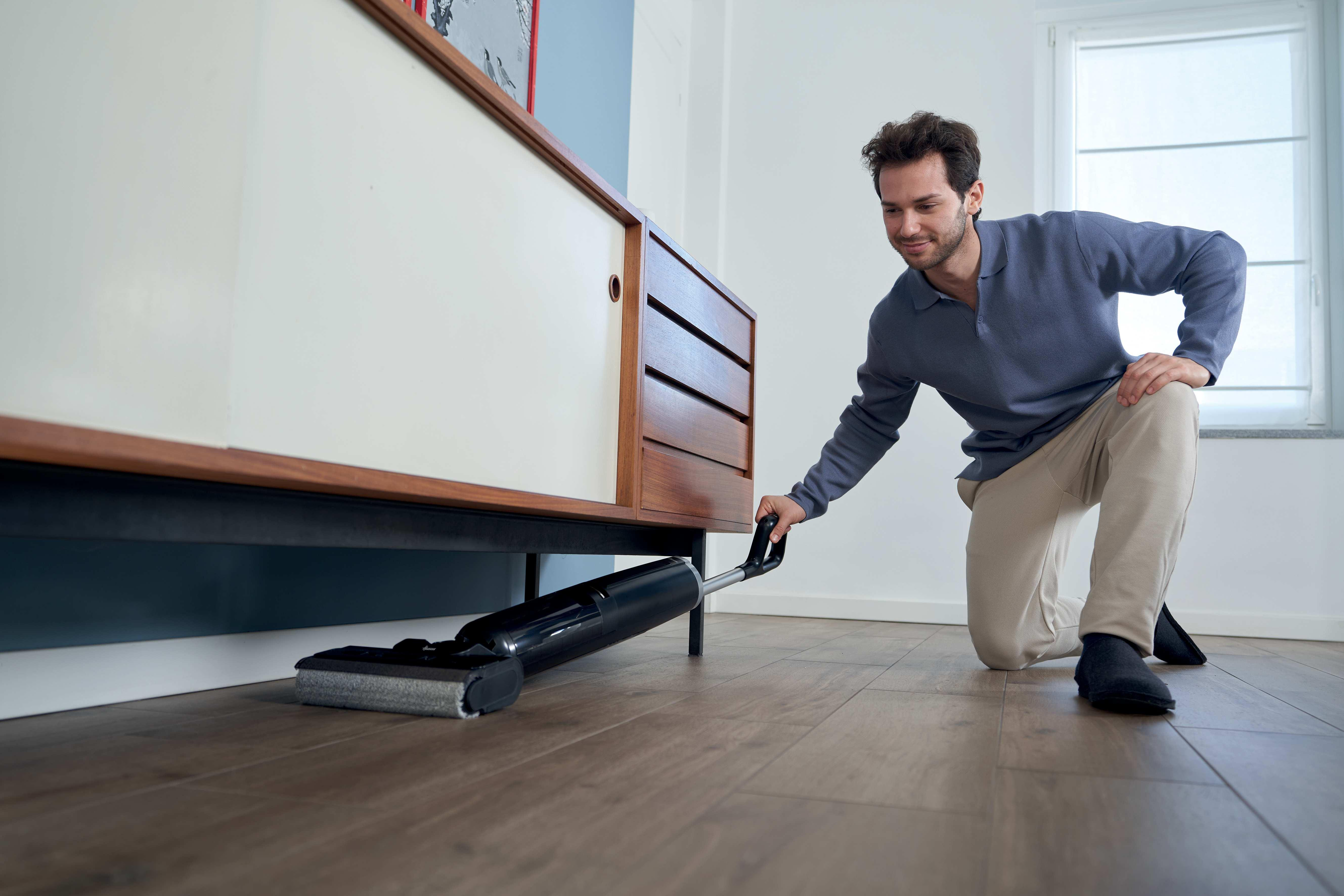 Un homme s'agenouille, passant l'aspirateur sous une armoire en bois avec un aspirateur noir.