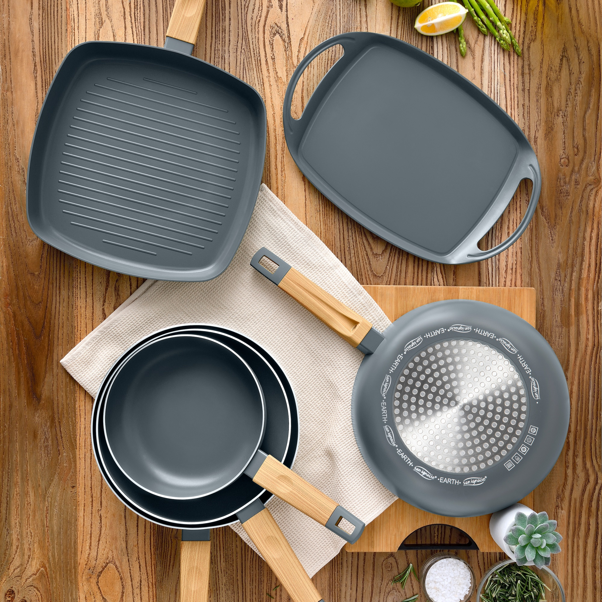 Utensilios de cocina grises con mangos de madera sobre mesa de madera, junto a rodajas de limón y espárragos.