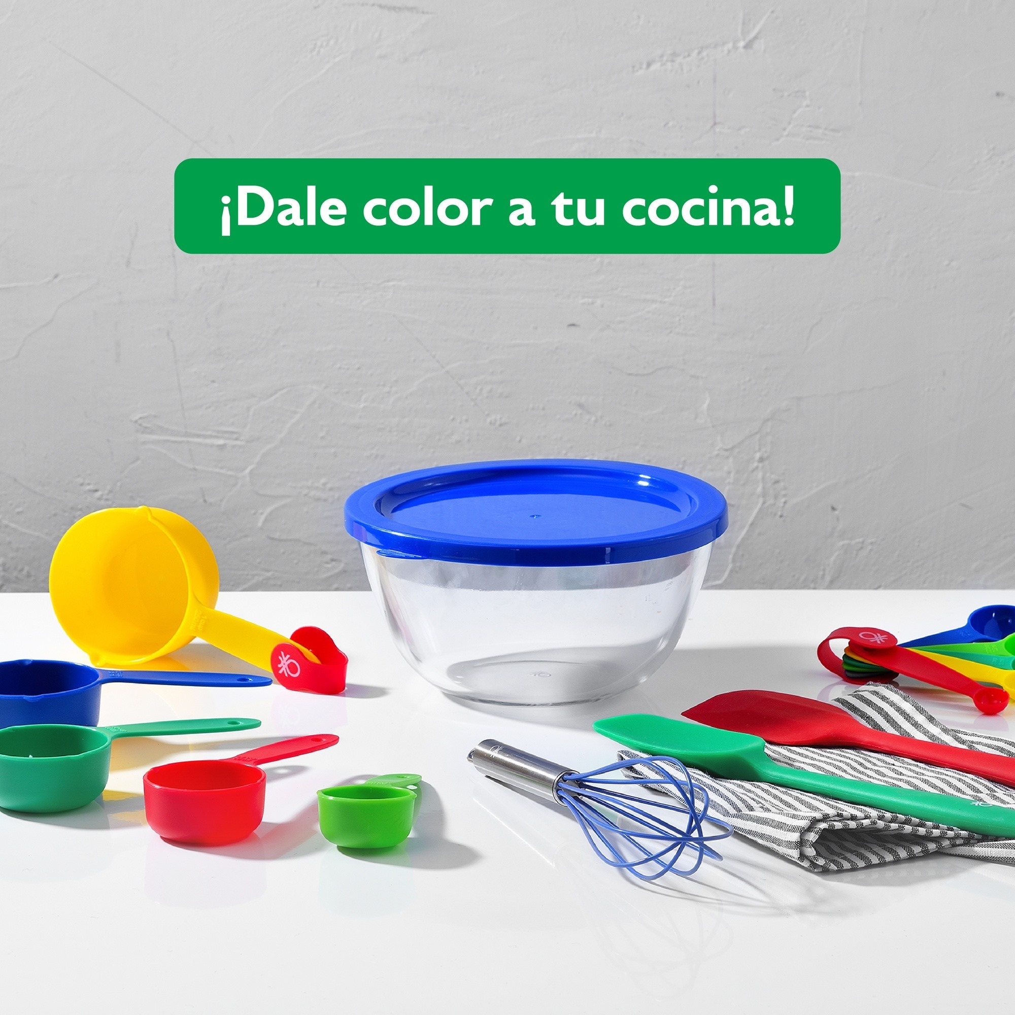 Utensilios de cocina coloridos, tazas medidoras, batidor y bol en mesa blanca.