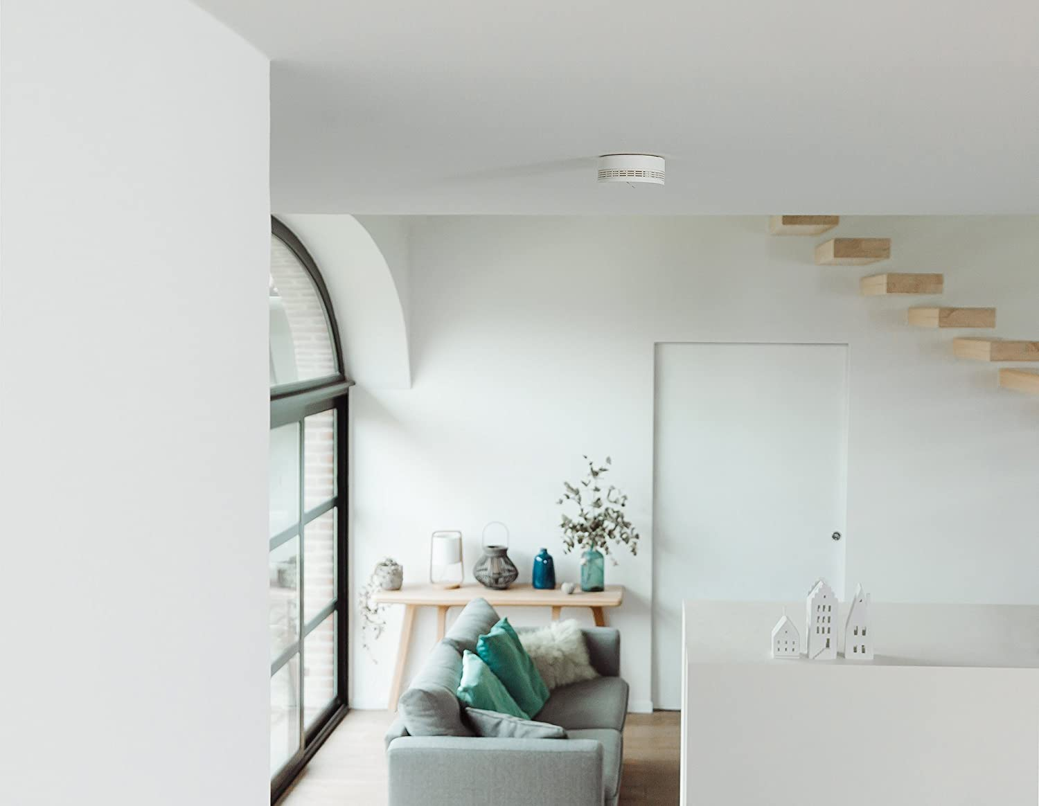A modern living room with a sofa, table, stairs, door, and smoke detector.