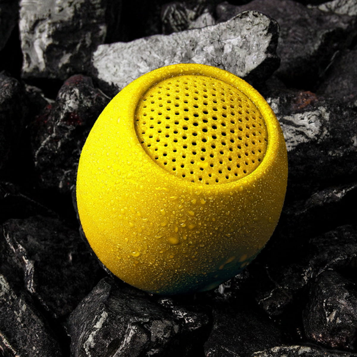 Un altoparlante giallo e rotondo con goccioline d'acqua poggia su pietre scure.