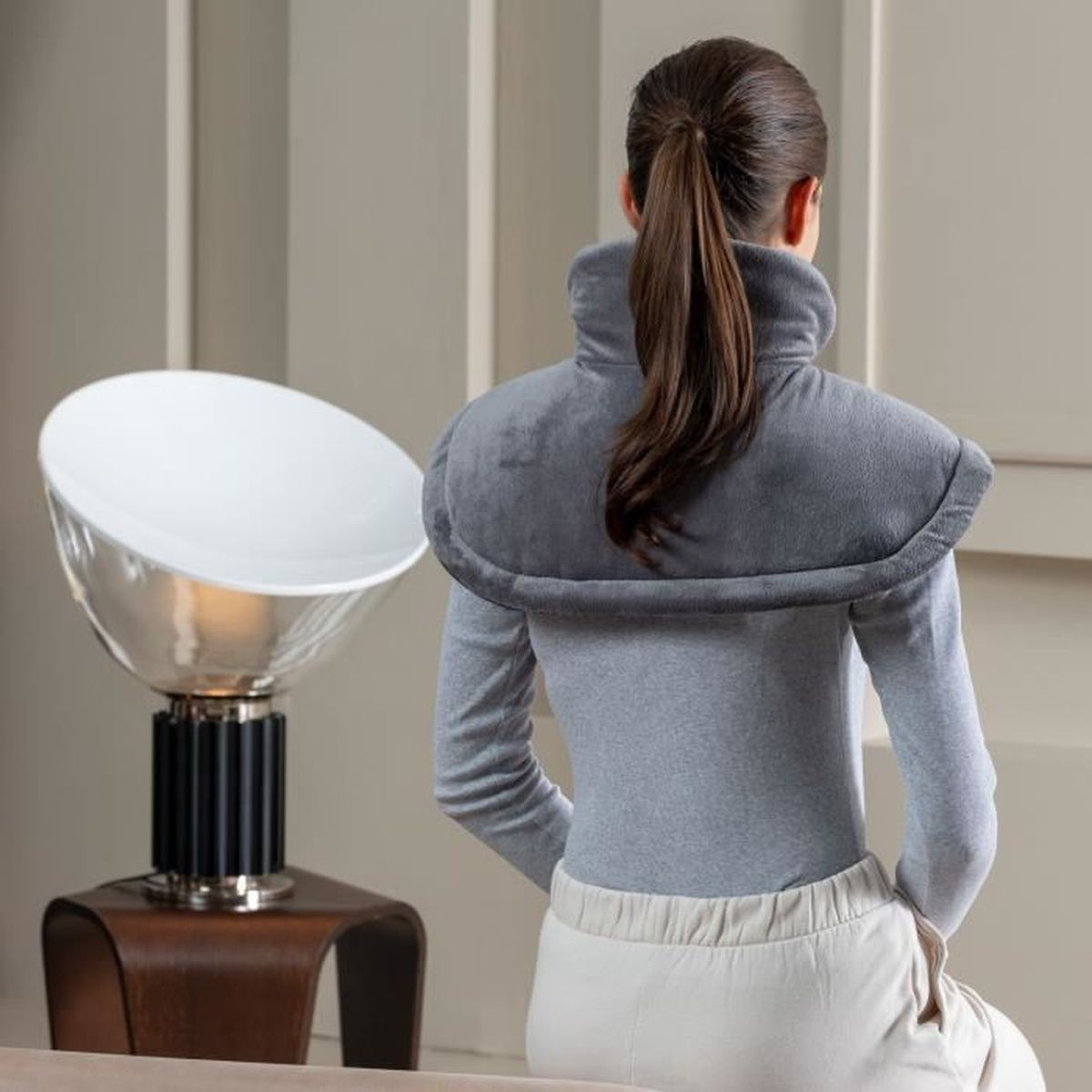 Mujer con chaleco gris y camisa gris, junto a una lámpara moderna.