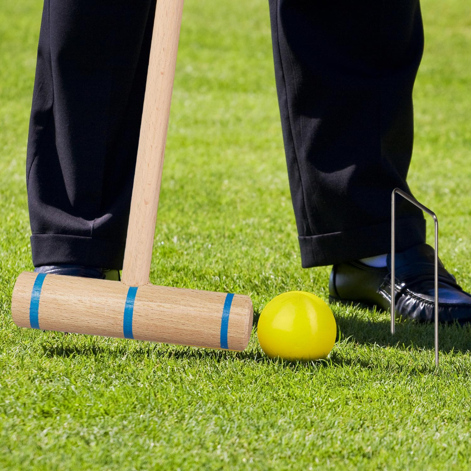 Una persona juega al croquet, golpeando una bola amarilla con un mazo de madera sobre césped.