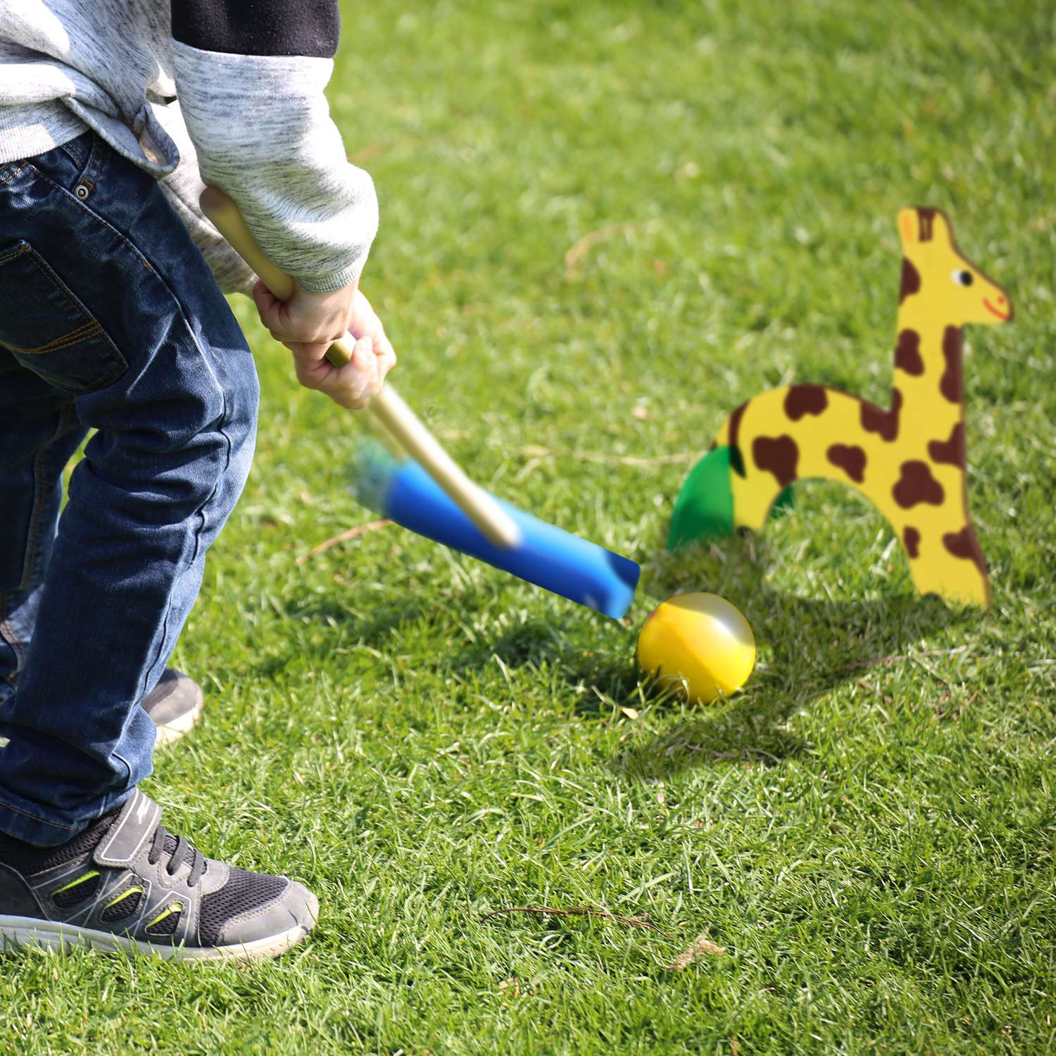 Niño blande bate contra balón amarillo y figura de jirafa en césped.