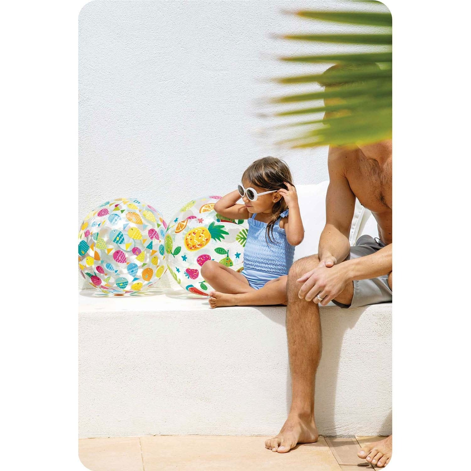 Un hombre y un niño sentados en un muro blanco junto a pelotas de playa coloridas.
