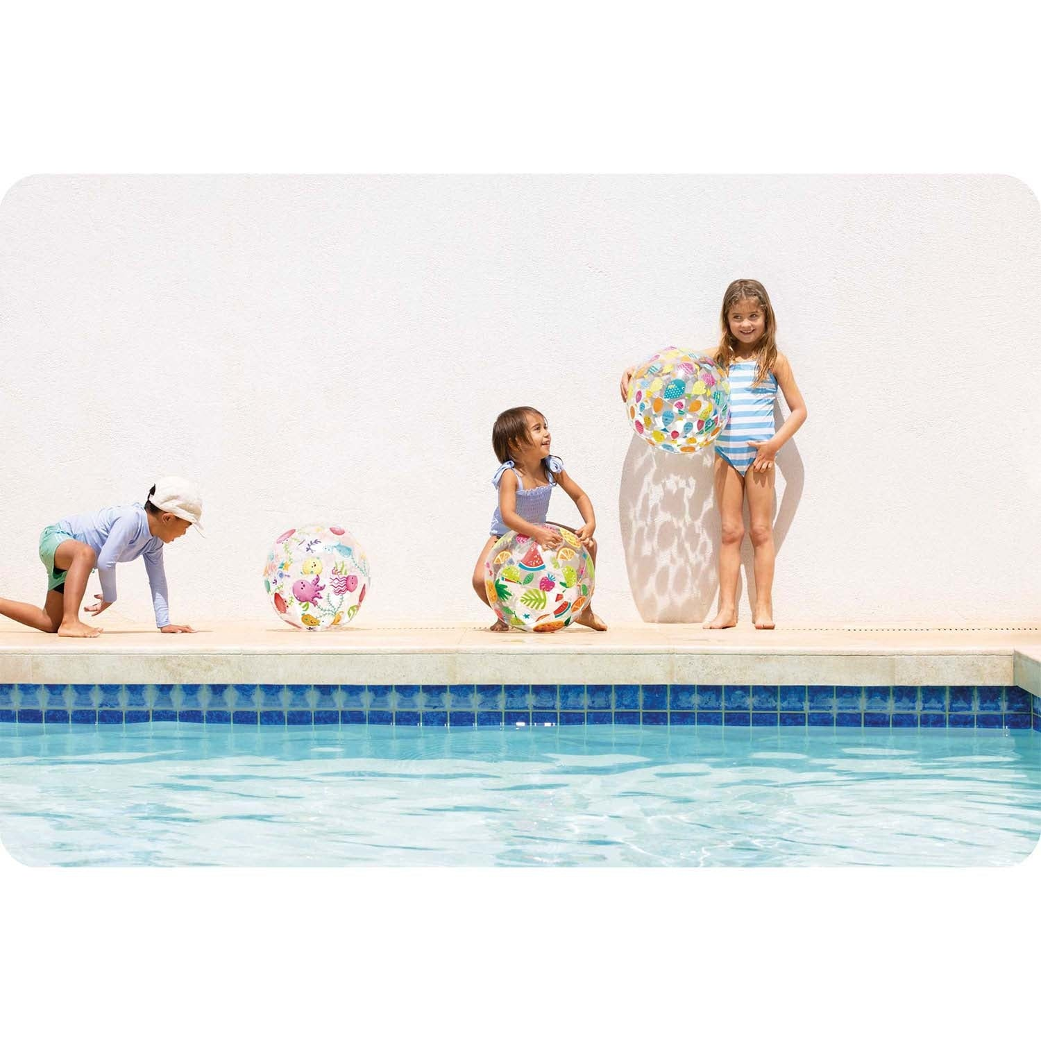 Tres niños junto a una piscina con pelotas coloridas frente a pared blanca.