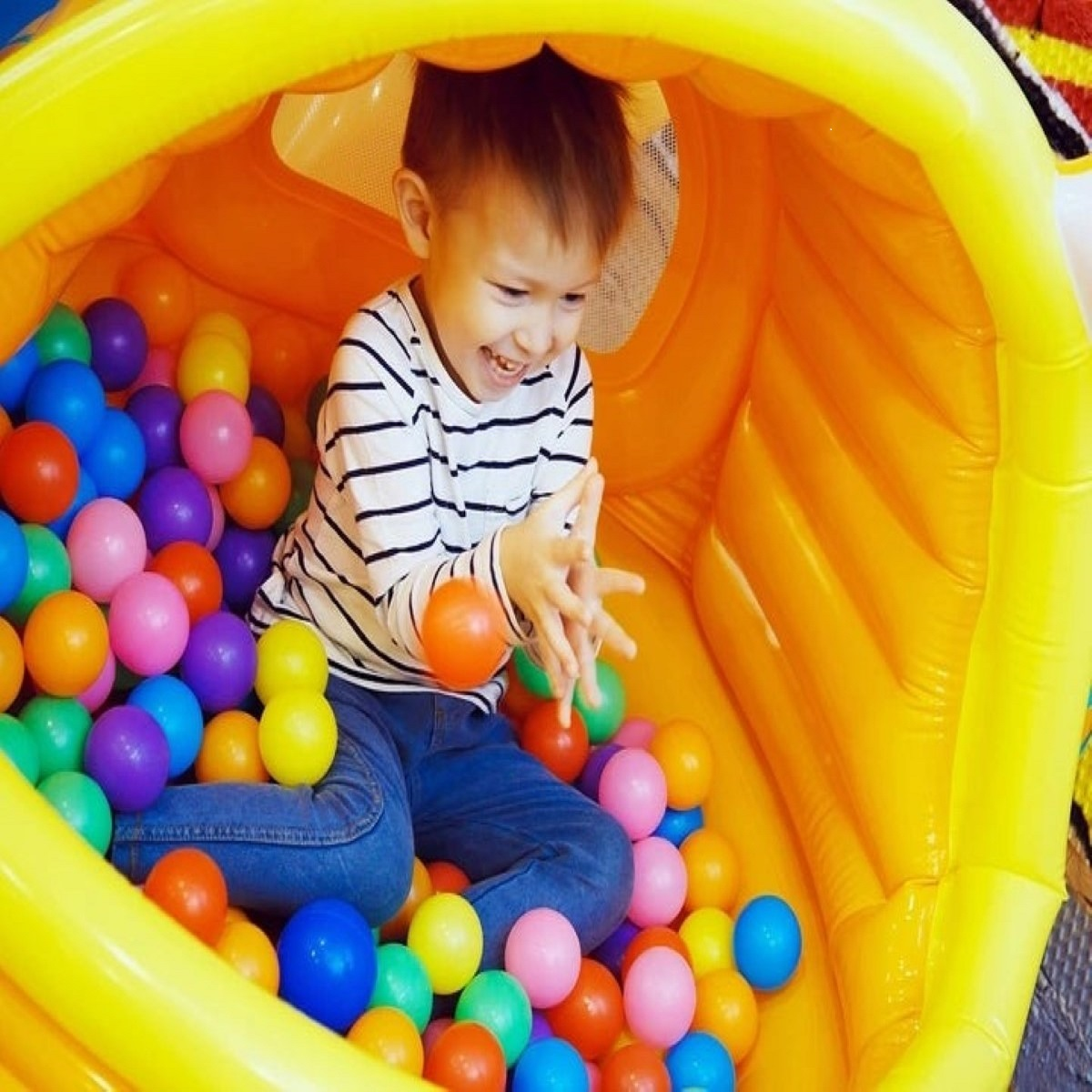 Un bambino con maglietta a righe gioca in una piscina di palline gialla con palline colorate.