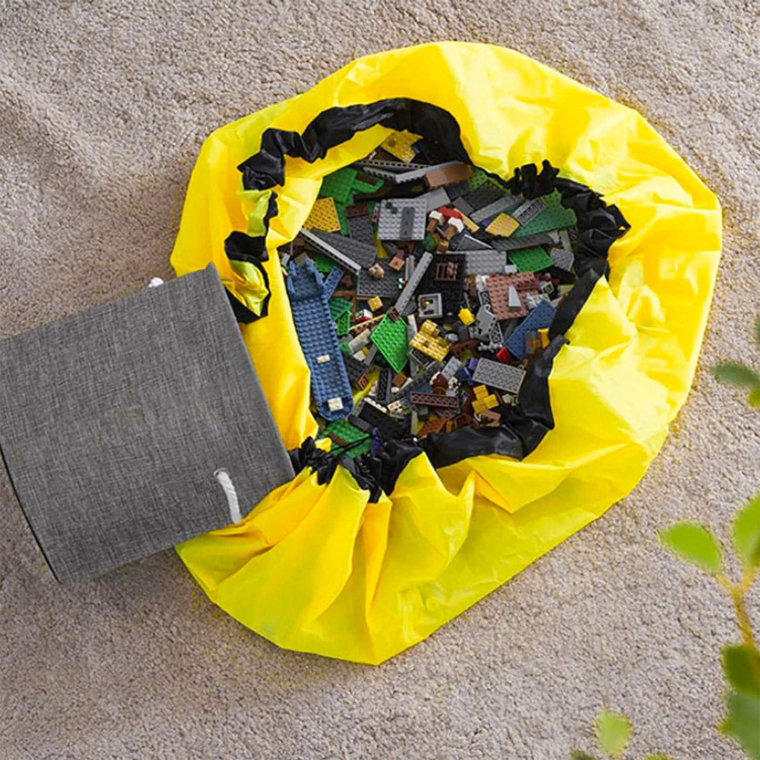 Una bolsa de juguetes amarilla llena de ladrillos Lego coloridos está en una alfombra. Una caja de almacenamiento gris está al lado.