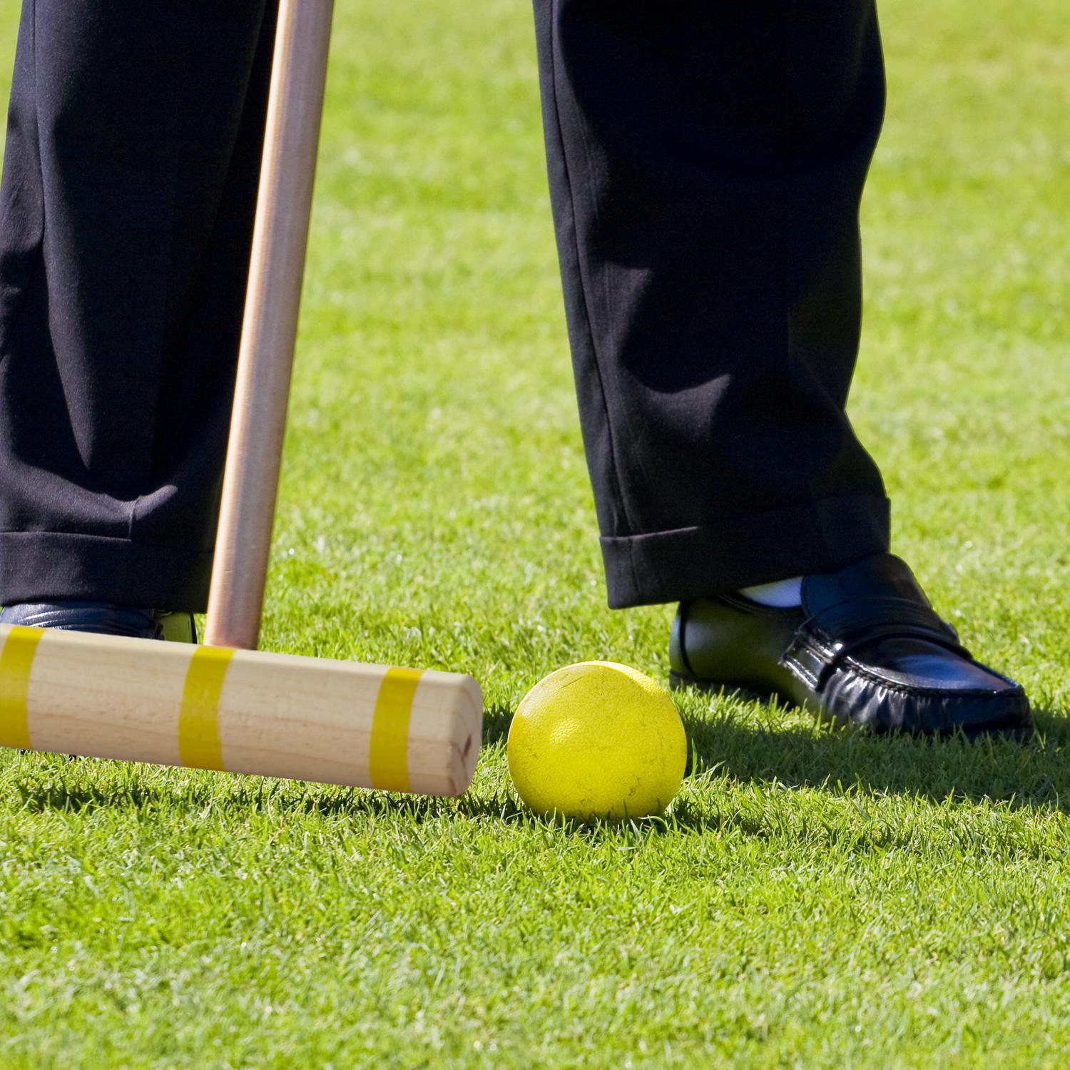 Un jugador con pantalones oscuros y zapatos se prepara para un golpe de croquet en césped verde.