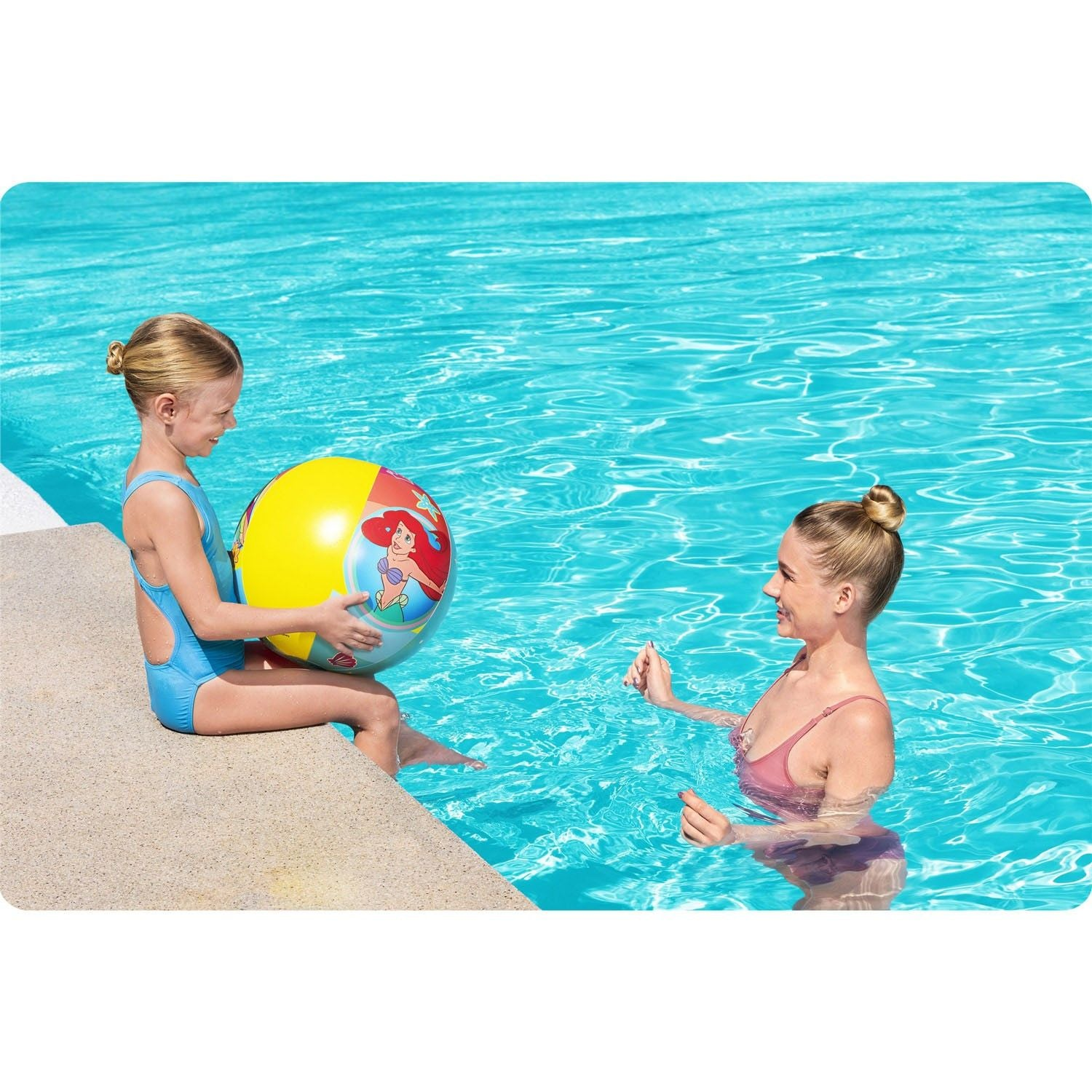 Una niña se sienta al borde de la piscina sosteniendo una pelota amarilla con Ariel. Una mujer está en el agua.