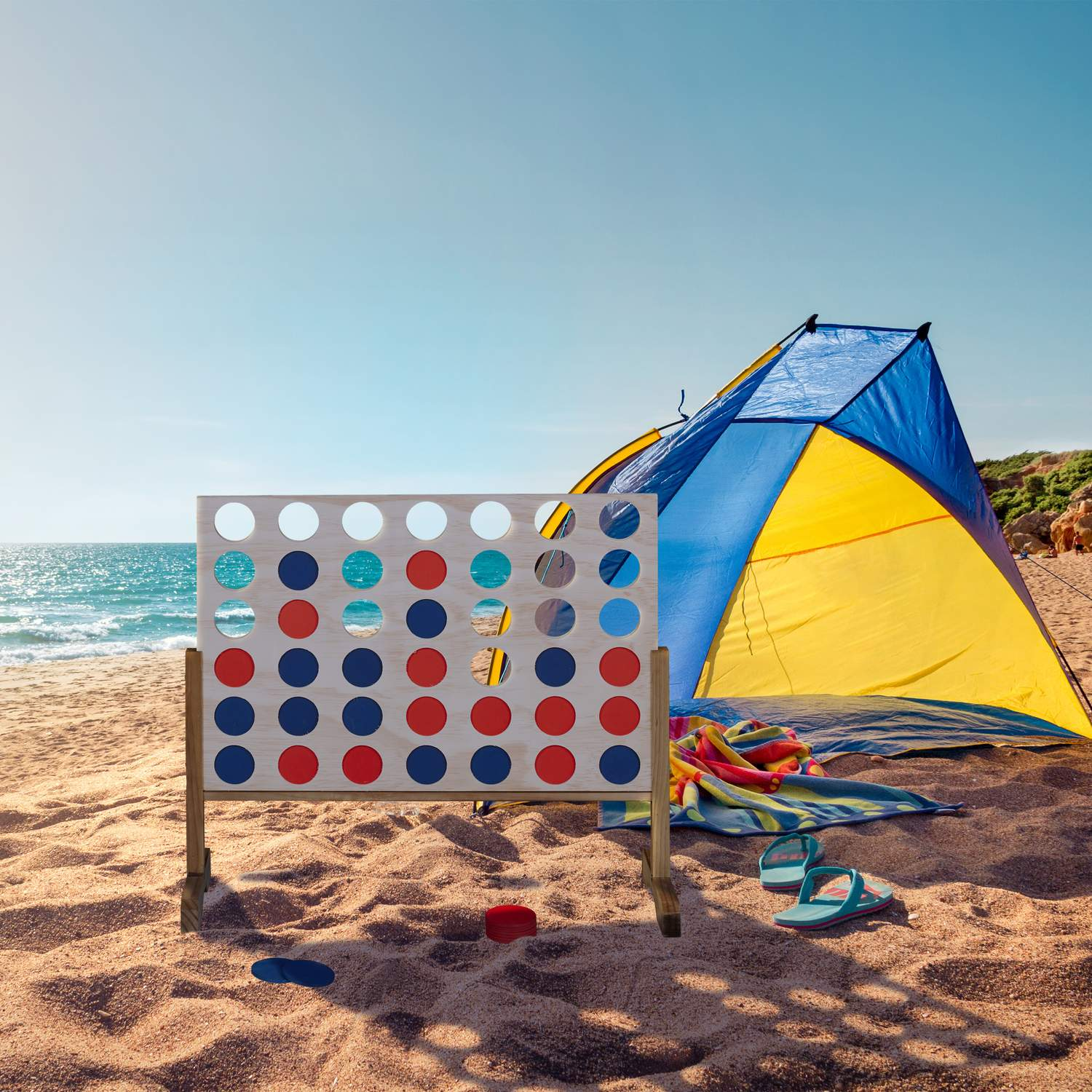 Escena de playa con juego gigante de conecta cuatro, tienda, toalla y chanclas.