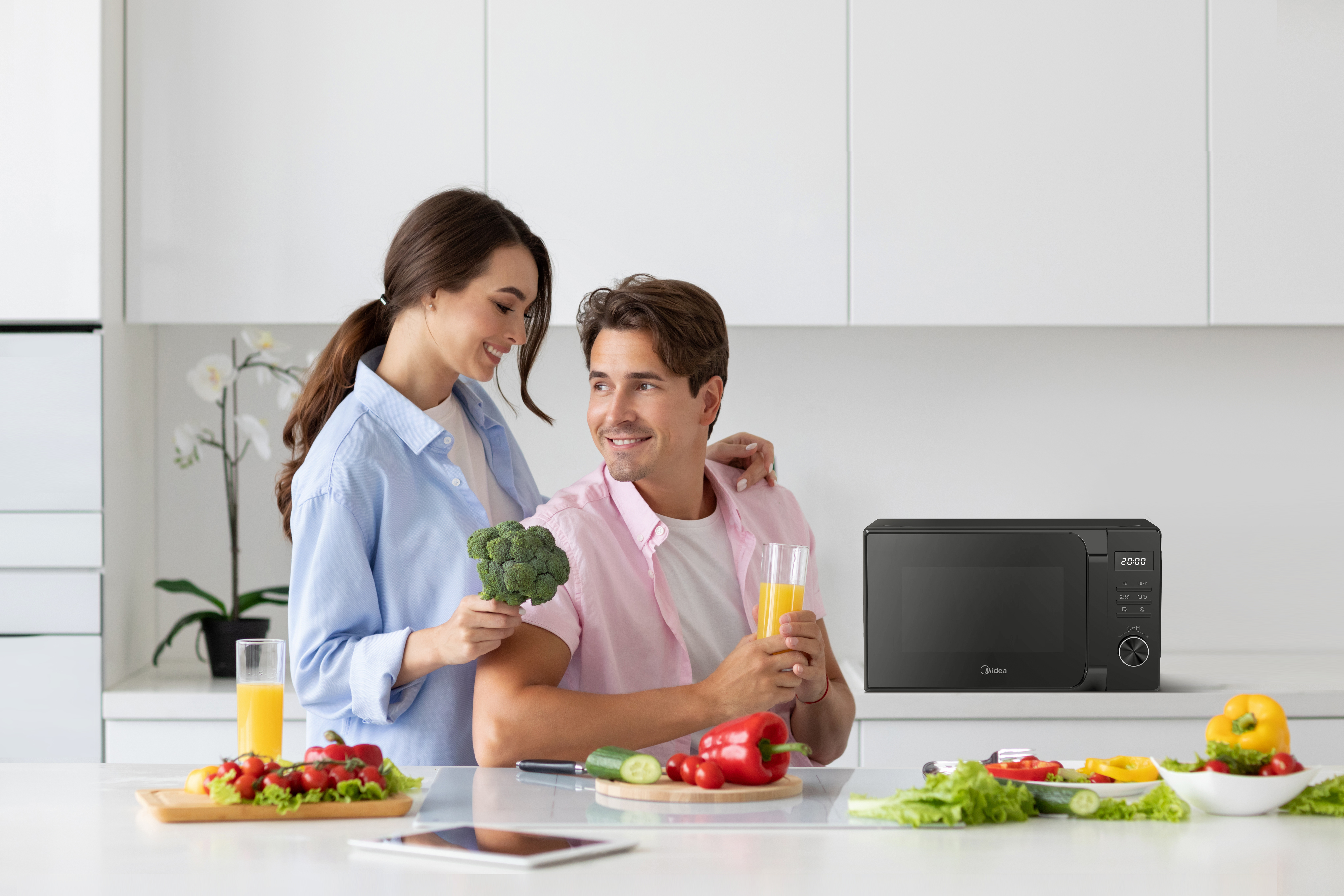 Una coppia in cucina, lei tiene i broccoli, lui beve succo. È presente un forno a microonde nero.