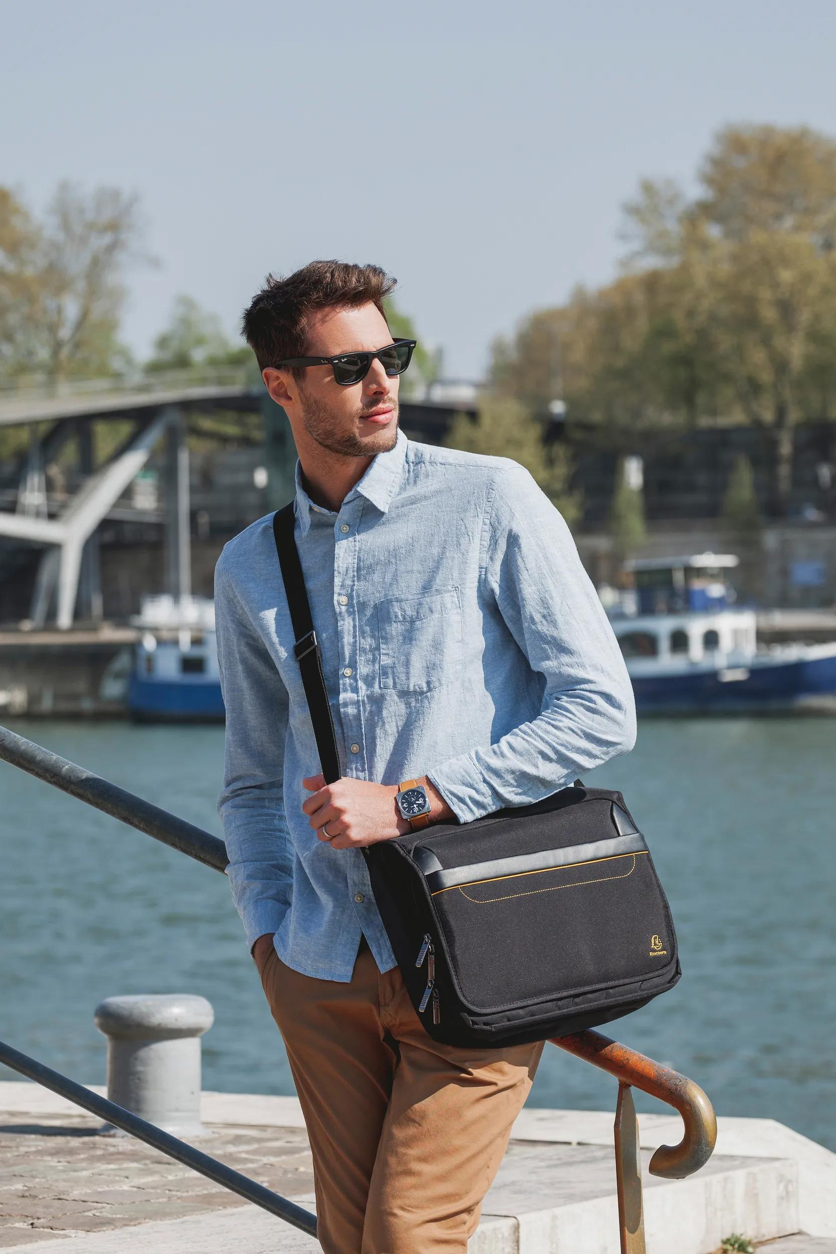 Hombre con bolso cruzado junto al agua, bote y árboles al fondo.