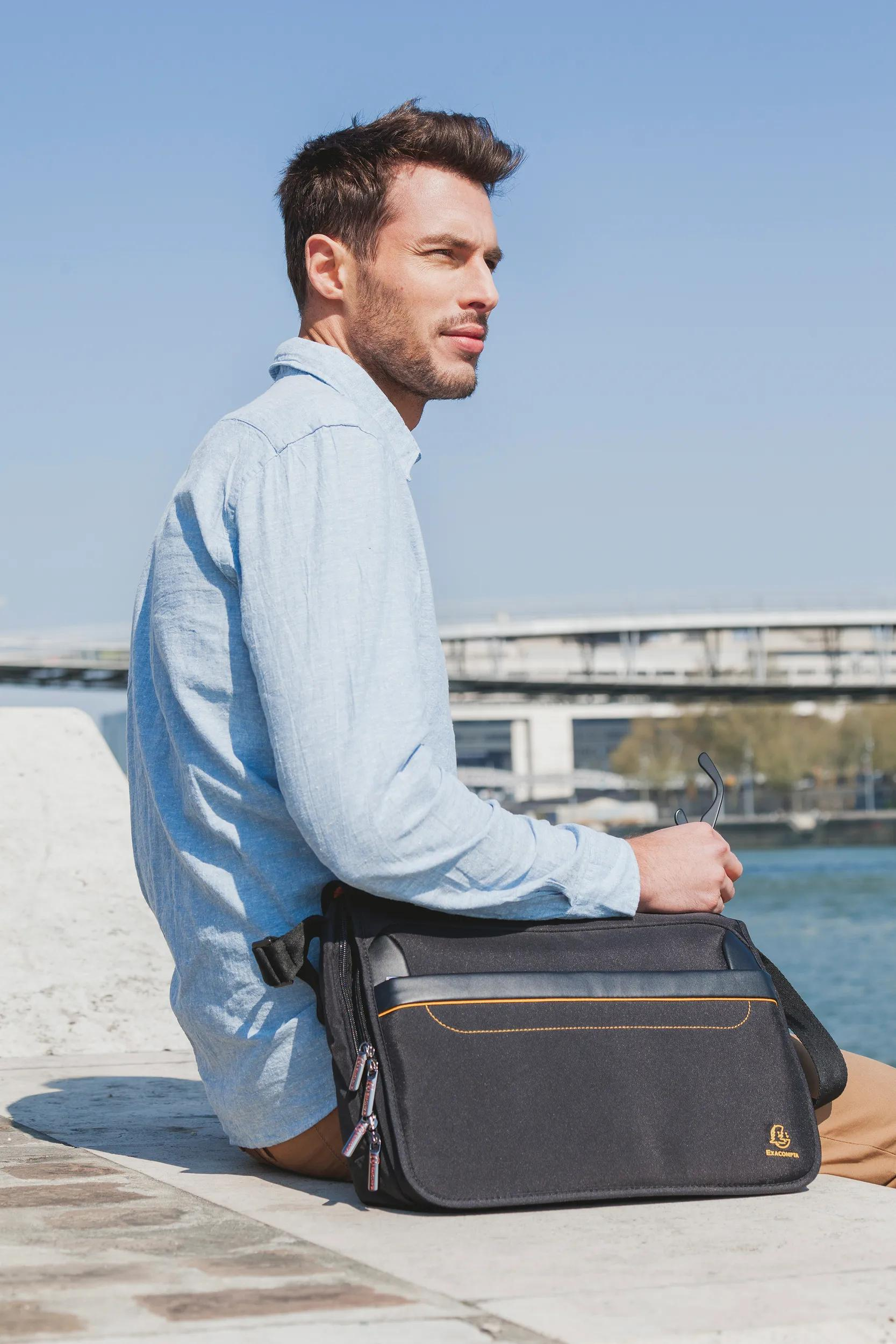 Hombre con camisa azul claro sentado junto al agua con bolso negro.