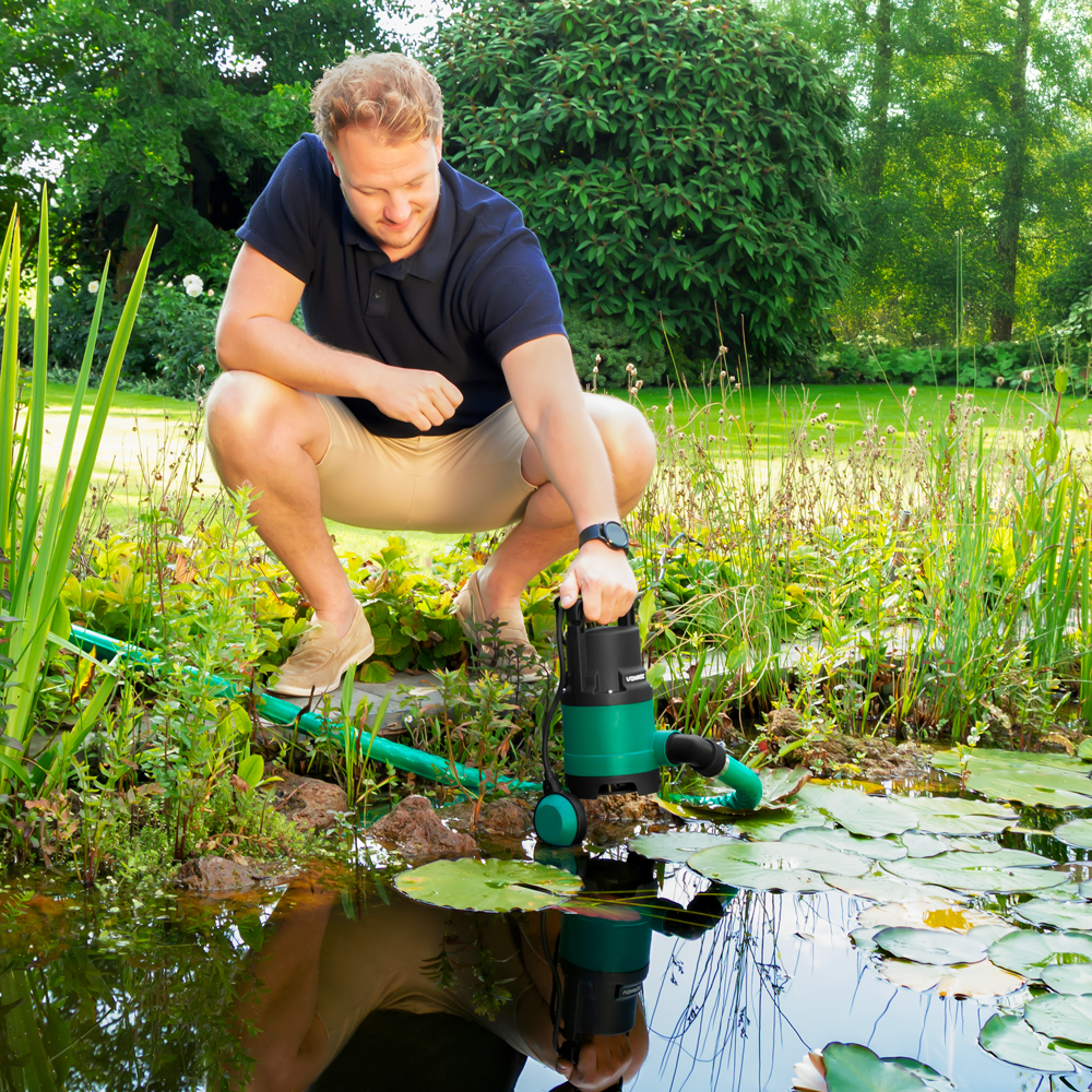 Man knielt bij een vijver en houdt een groene pomp met zwart handvat vast.