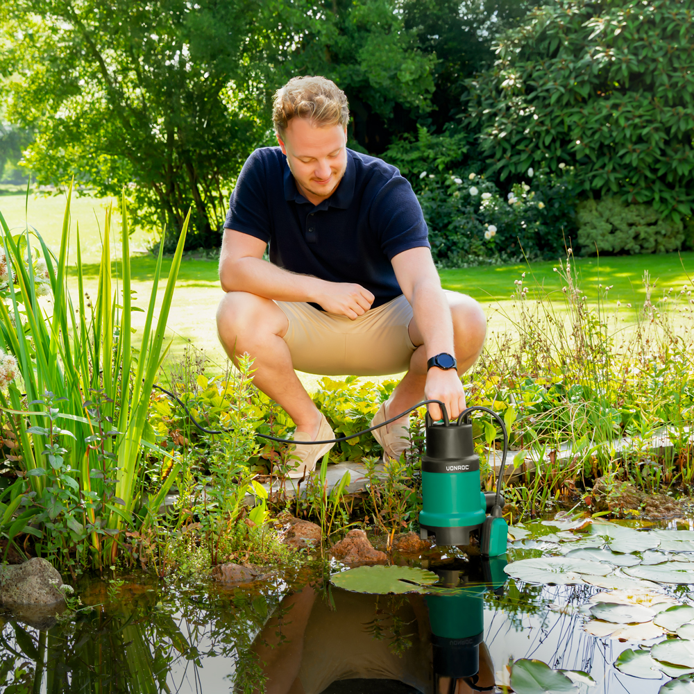 Man in een tuin plaatst een groene pomp in een vijver vol waterplanten.